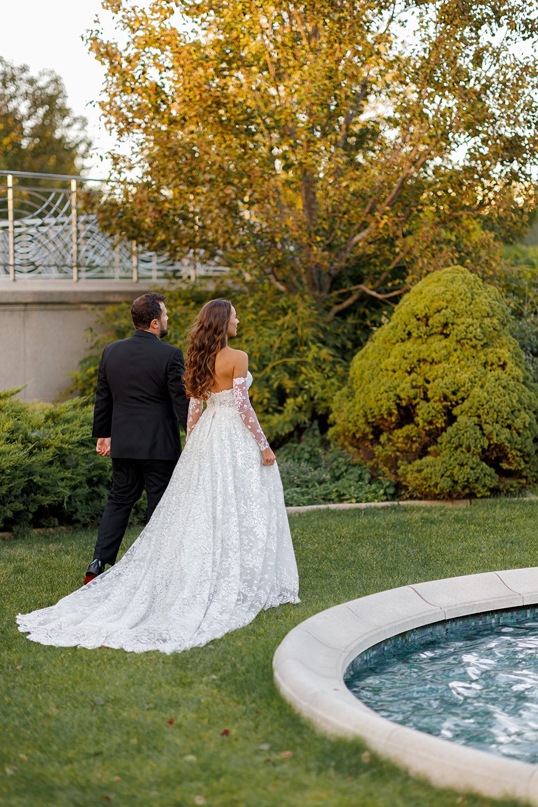 Bride in lace off-shoulder gown with long train walks with groom in black tuxedo through elegant garden.