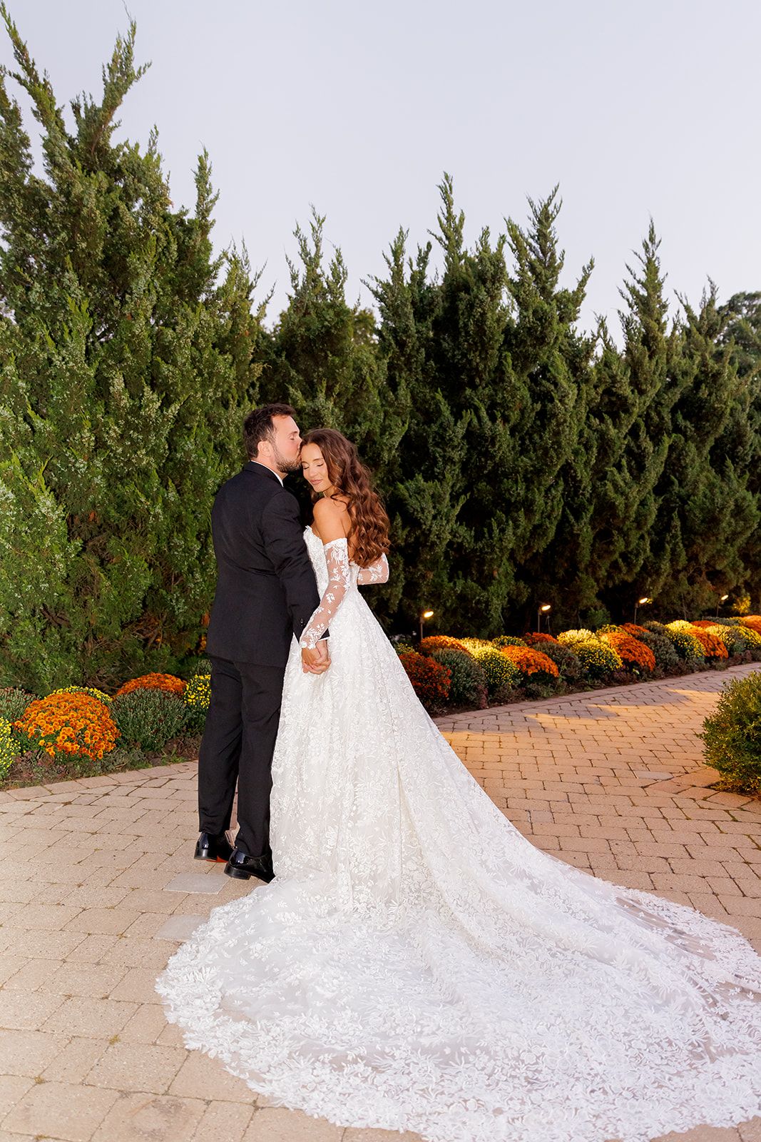 Newlyweds share a tender kiss on a brick path, bride in a stunning lace gown with long train, flanked by fall flowers.