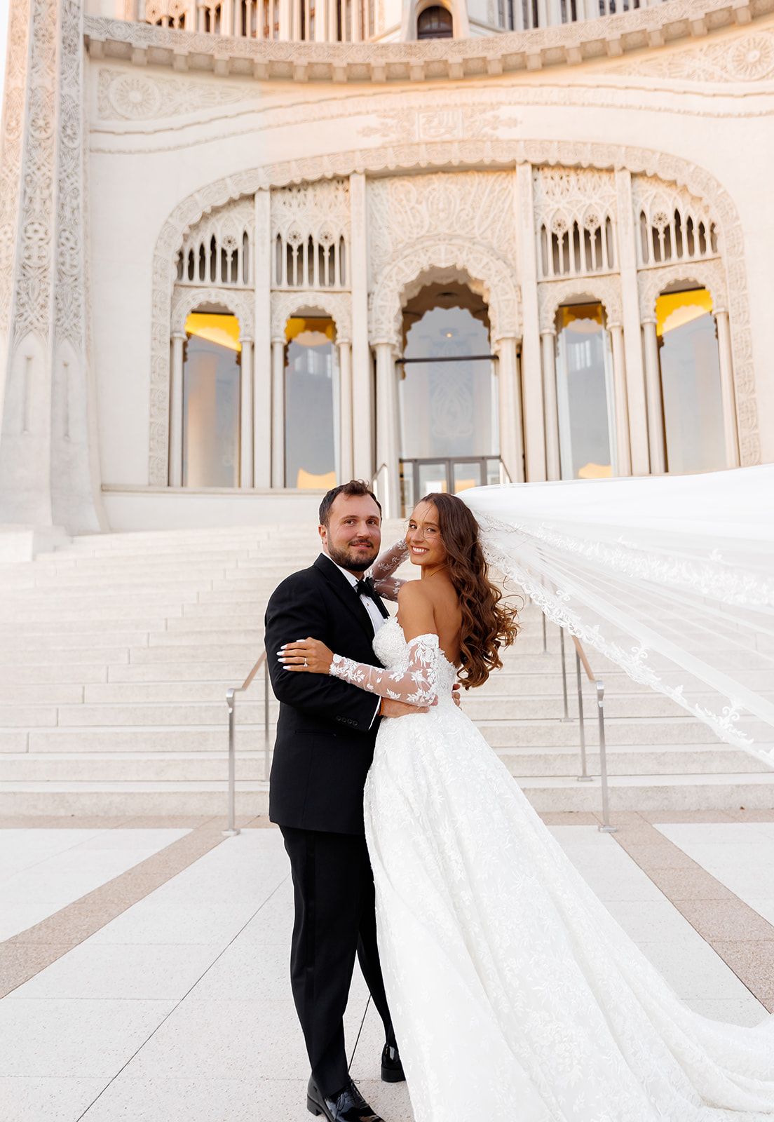 Smiling couple poses before ornate white temple as bride's cathedral veil billows in the wind.