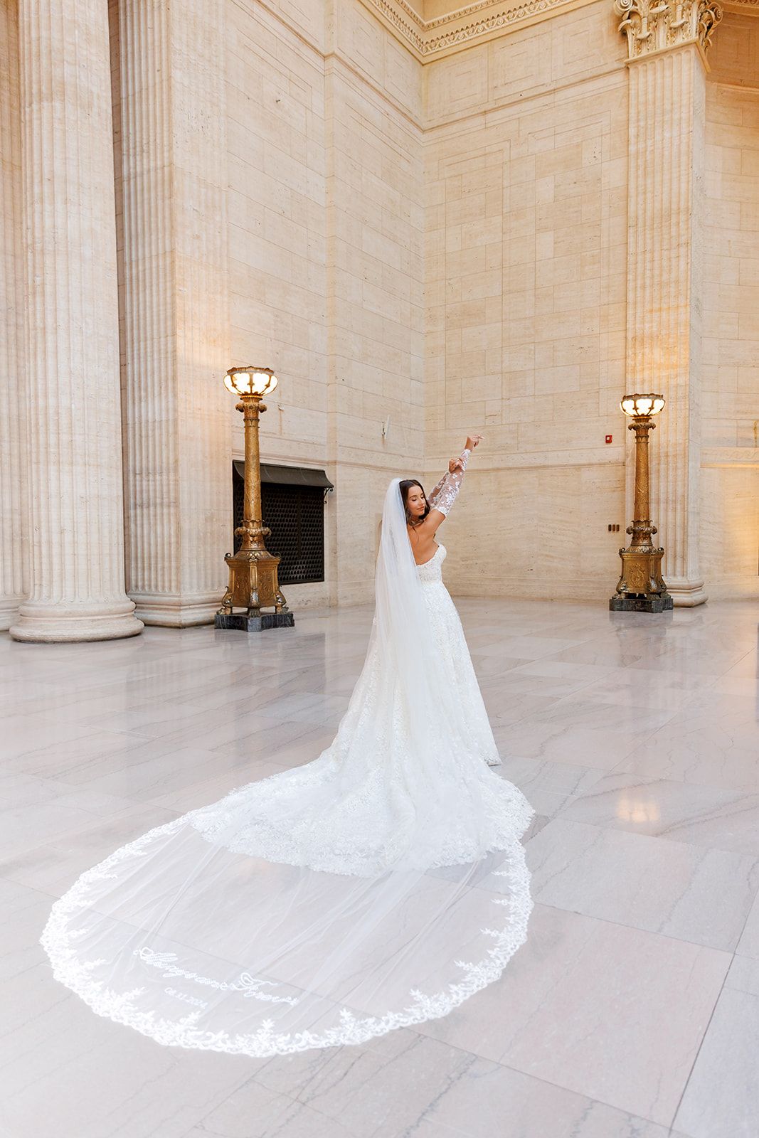 Bride in white gown with long lace-trimmed cathedral veil poses joyfully in grand marble hall with tall columns.