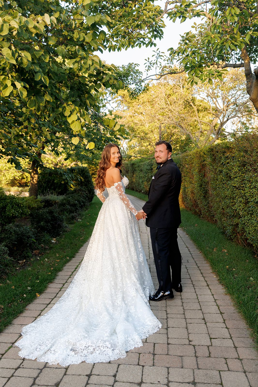 Bride in lace gown and groom in black tuxedo holding hands on garden pathway surrounded by green trees.
