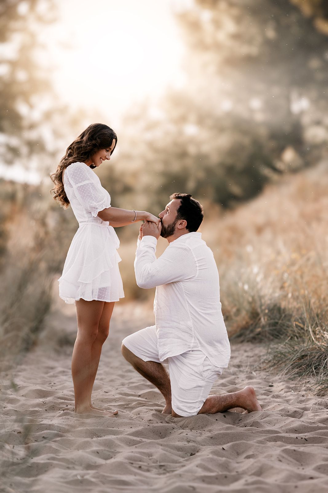 Man in white kneels on sandy beach kissing woman's hand during golden hour proposal.
