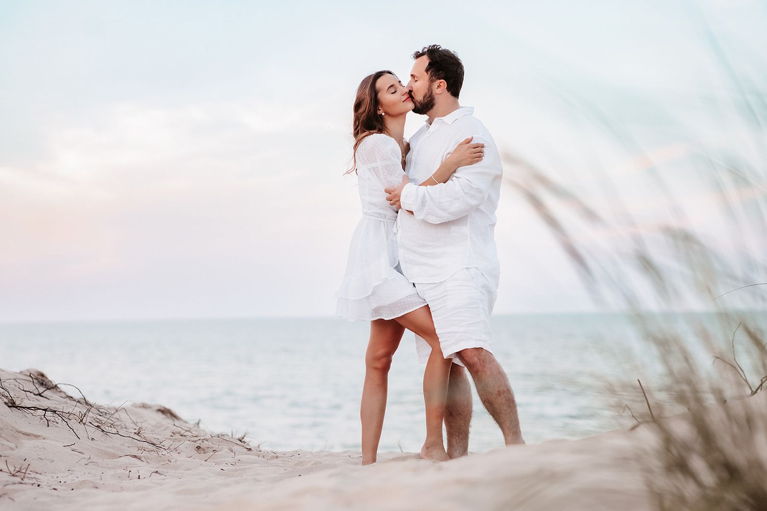 Couple in white outfits kissing on a sandy beach with sea grass and calm ocean behind them.