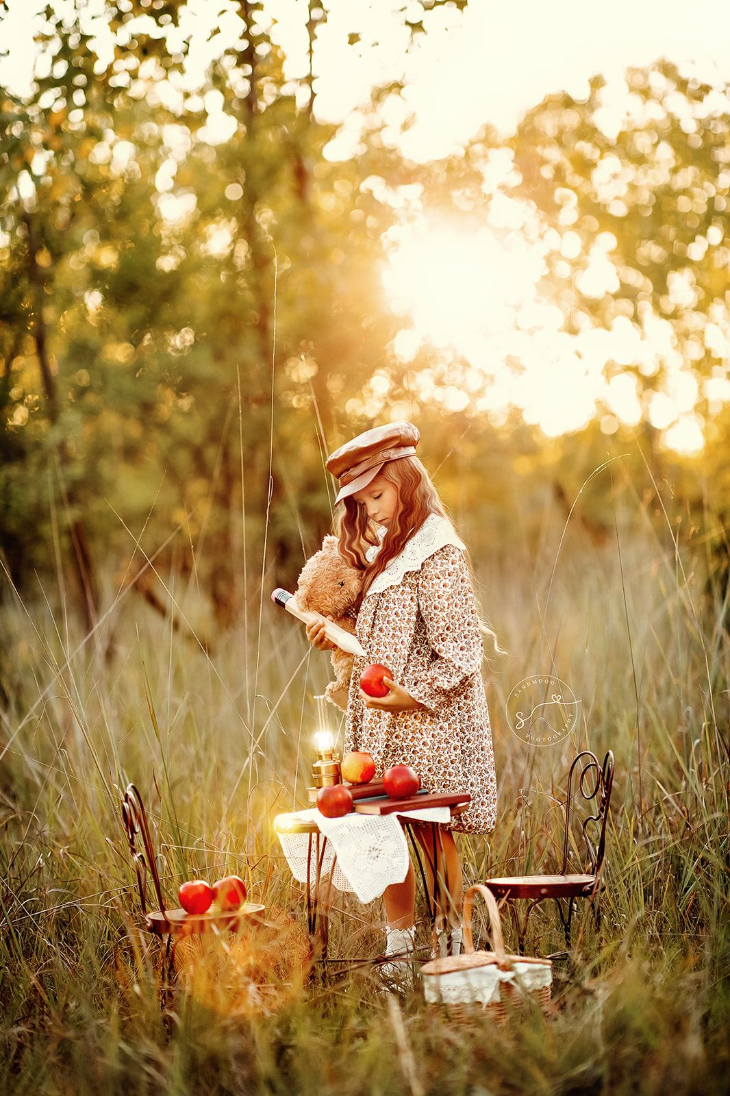 A young girl in a floral dress and hat holds a teddy bear at a golden sunset picnic in a meadow.