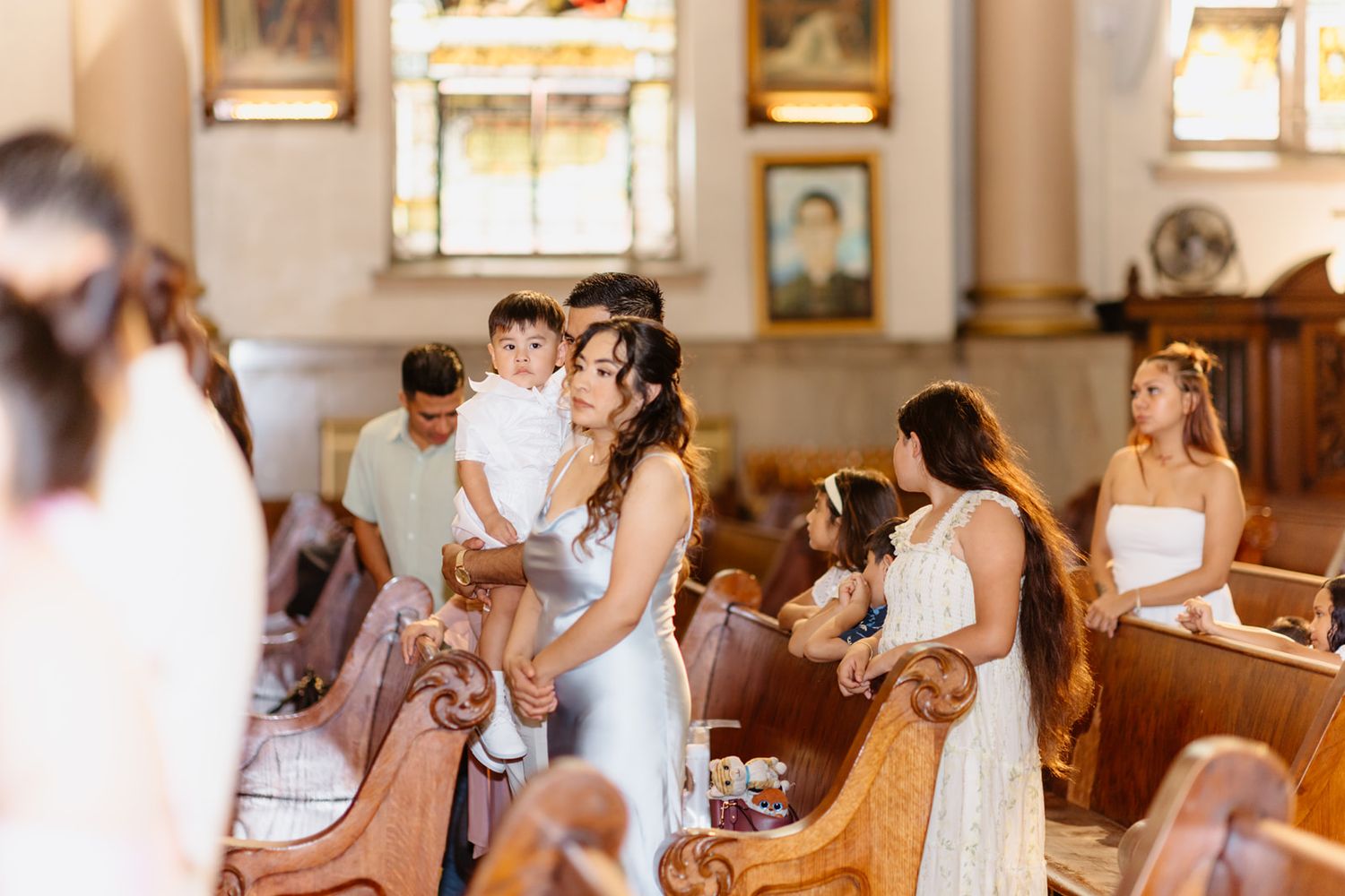 Woman in white dress holds toddler while guests gather in church pews during a baptism ceremony.