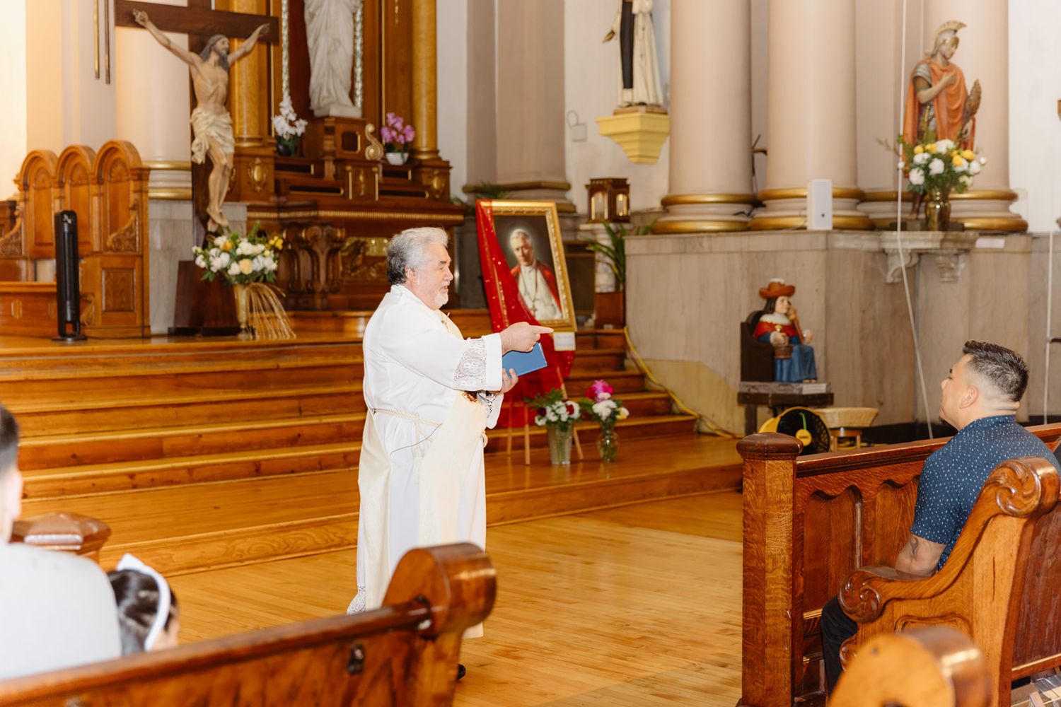 Priest holds religious icon during Catholic church ceremony with wooden pews and ornate altar in background.