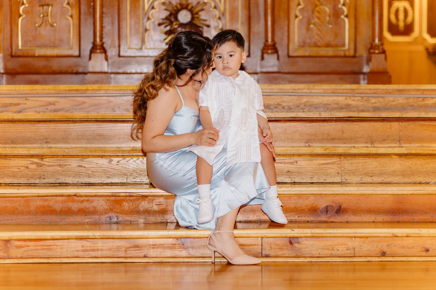 A mother in a blue dress sits on wooden church steps holding her baby in a white christening gown.