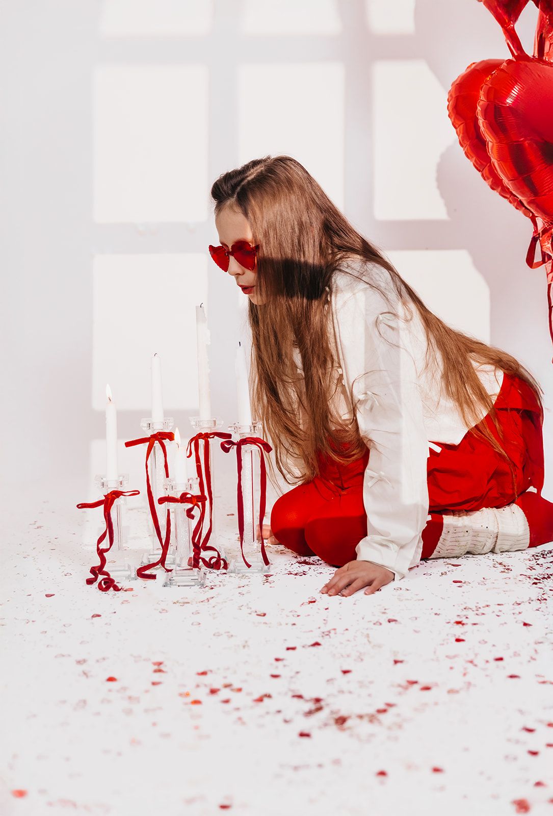 Girl in red pants and heart sunglasses kneeling near candles and red heart balloons on confetti-covered floor.