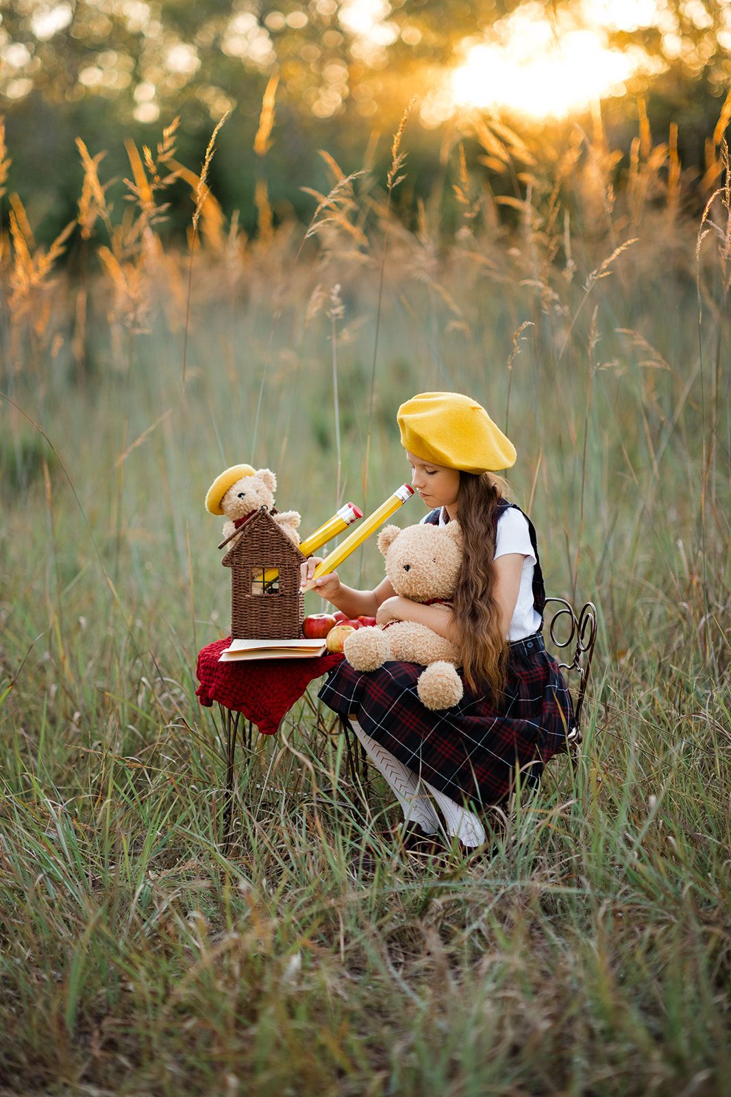 Girl in yellow beret sits in golden field with teddy bears at a small table during sunset.