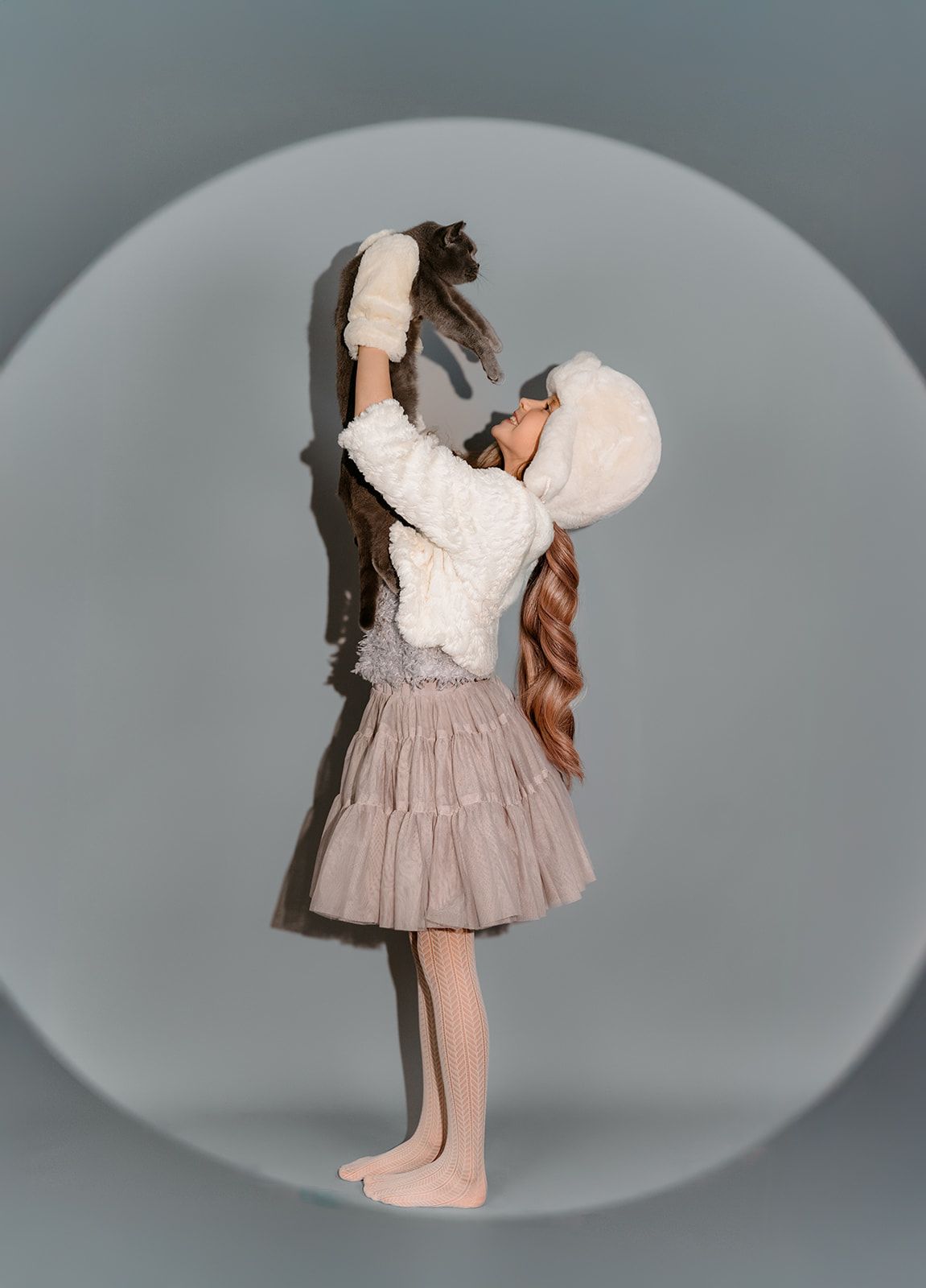 A barefoot girl in a white fluffy jacket and tulle skirt holds a cat above her head on a gray background.