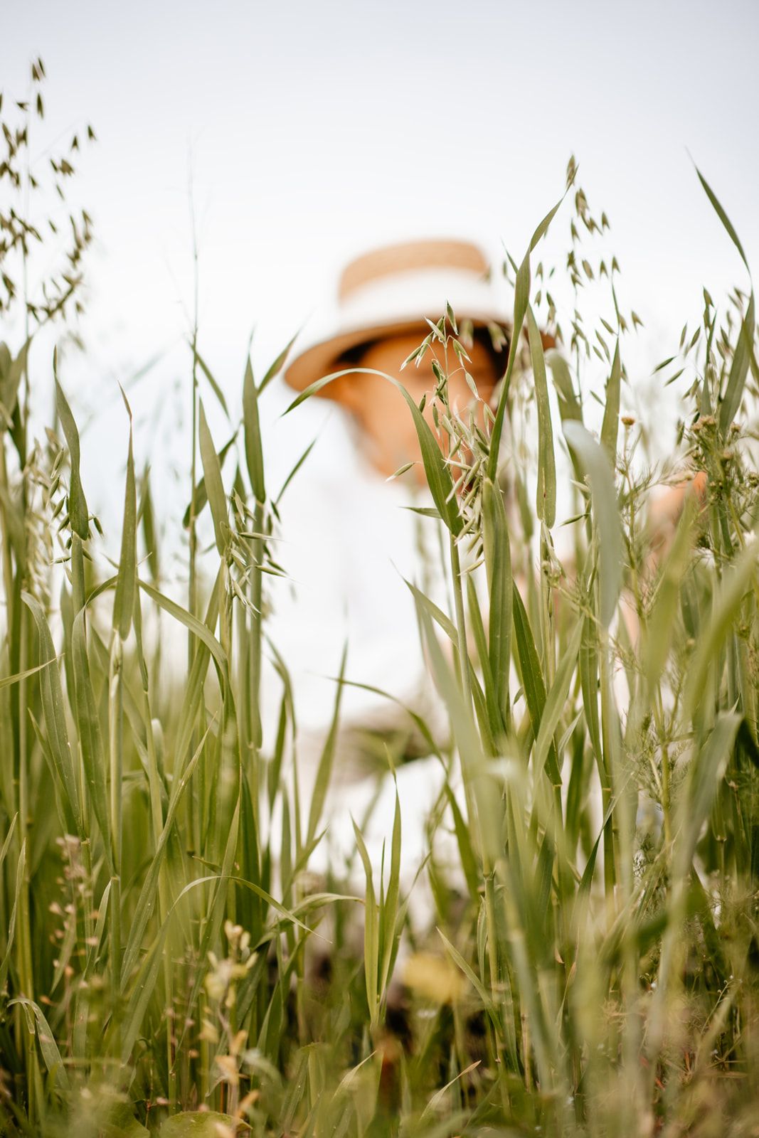 A woman in a white outfit and straw hat sits among tall green grass in a field.