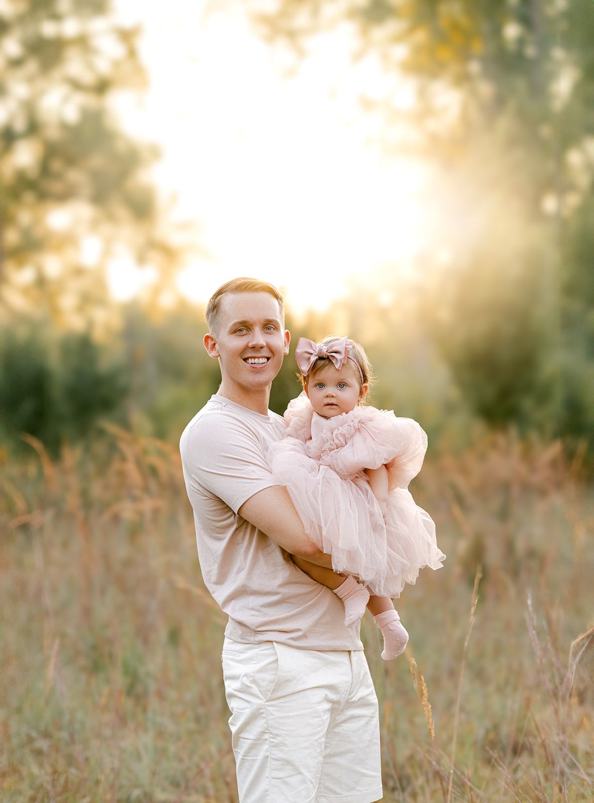 A smiling man holds a baby girl in a pink tutu dress outdoors in a golden sunlit field.