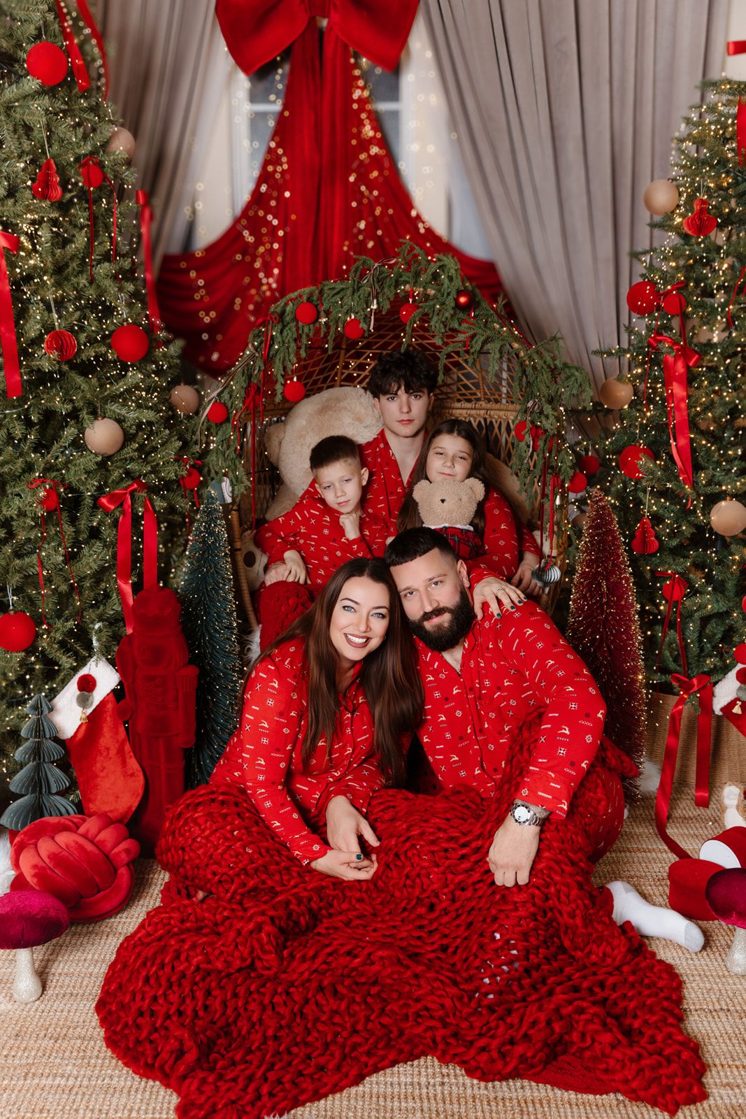Family in matching red pajamas posing together by decorated Christmas trees with red bows and ornaments.