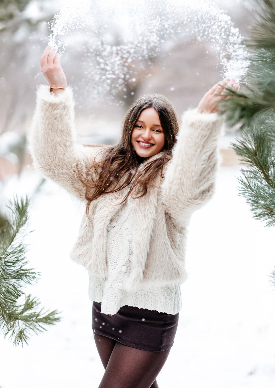 Young woman in white fluffy jacket tosses snow joyfully among pine trees in a winter wonderland setting.