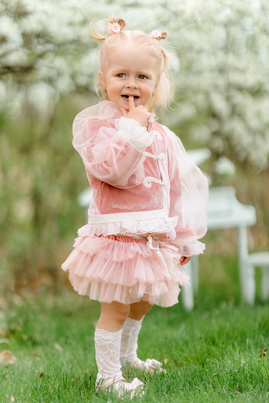 Adorable blonde toddler girl in pink tulle dress and white socks posing outdoors near blooming spring trees.