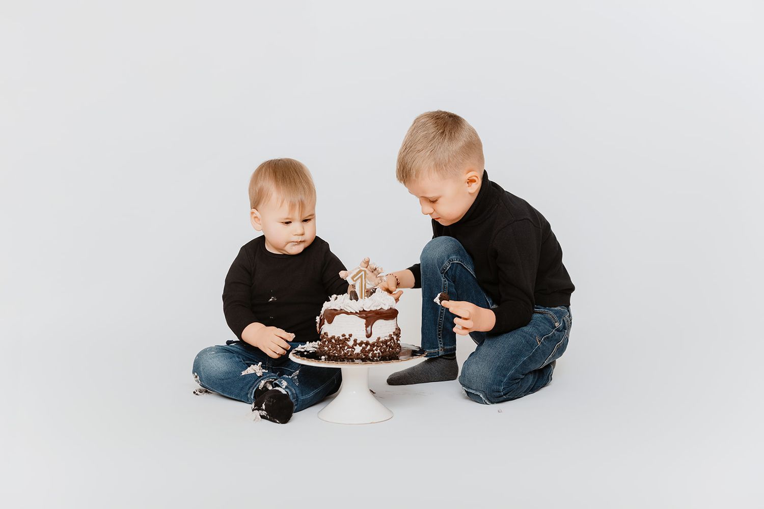 Two blonde boys in black tops sit on floor exploring decorated birthday cake together.