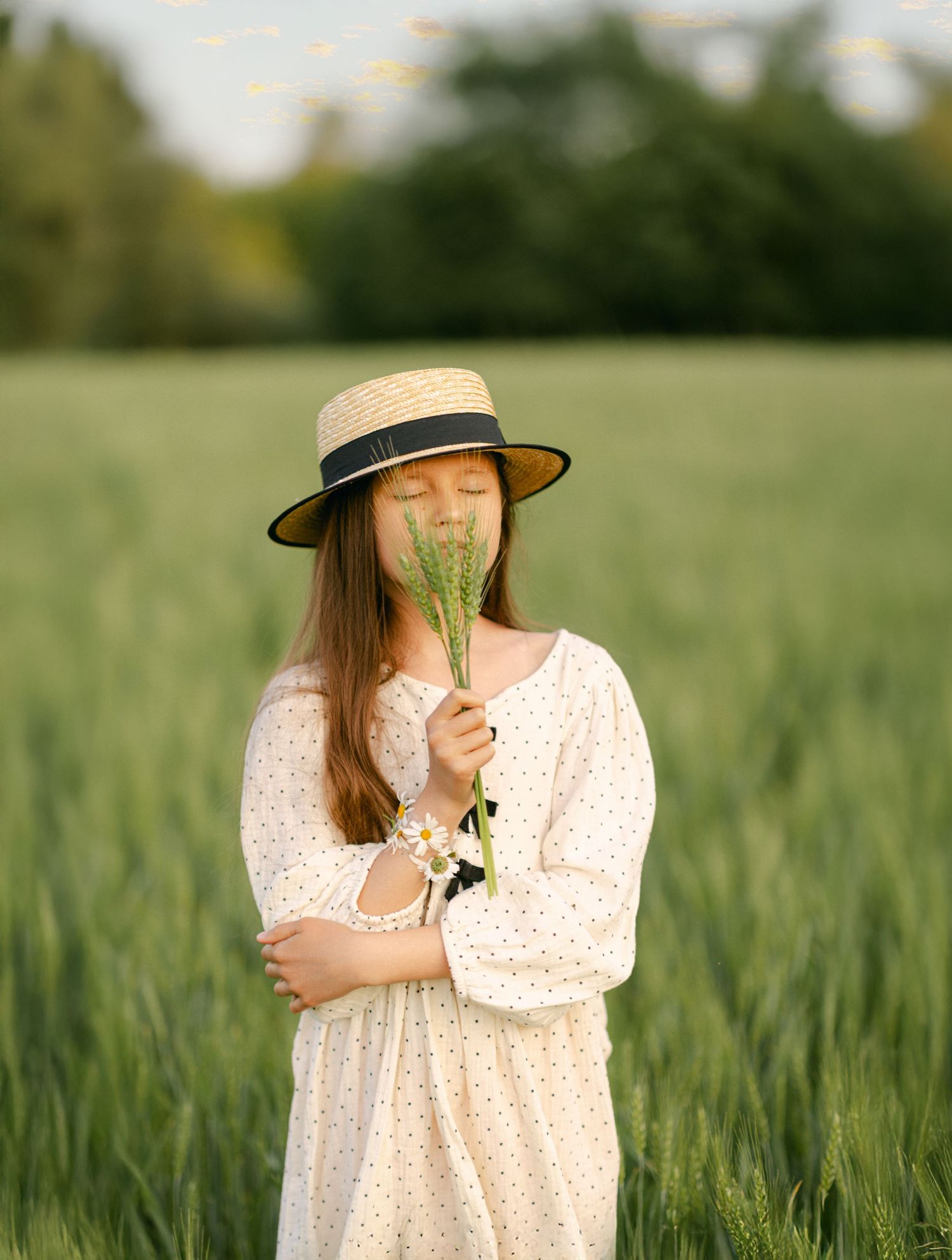 A young girl in a white polka dot dress and straw hat smells wildflowers in a lush green field.