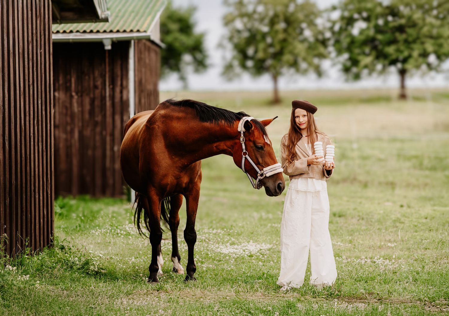 Young woman in white dress and hat stands beside a chestnut horse near a barn in a green rural field.