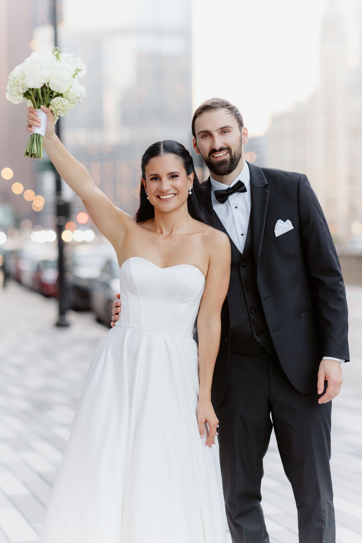 Bride in strapless white gown raises bouquet while groom in black tuxedo smiles on city street.
