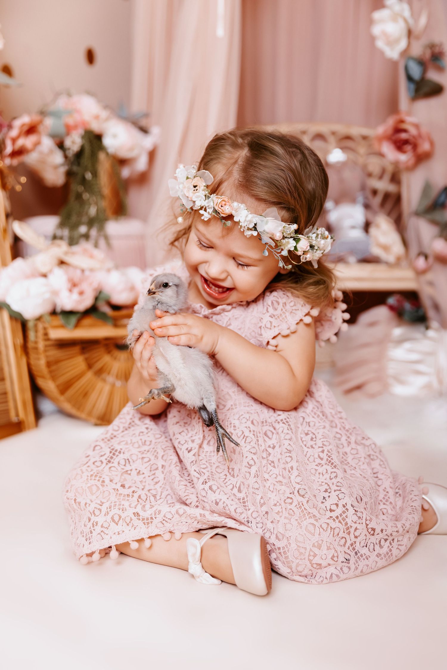 A joyful little girl in a pink lace dress and floral crown laughs while holding a small gray bunny.