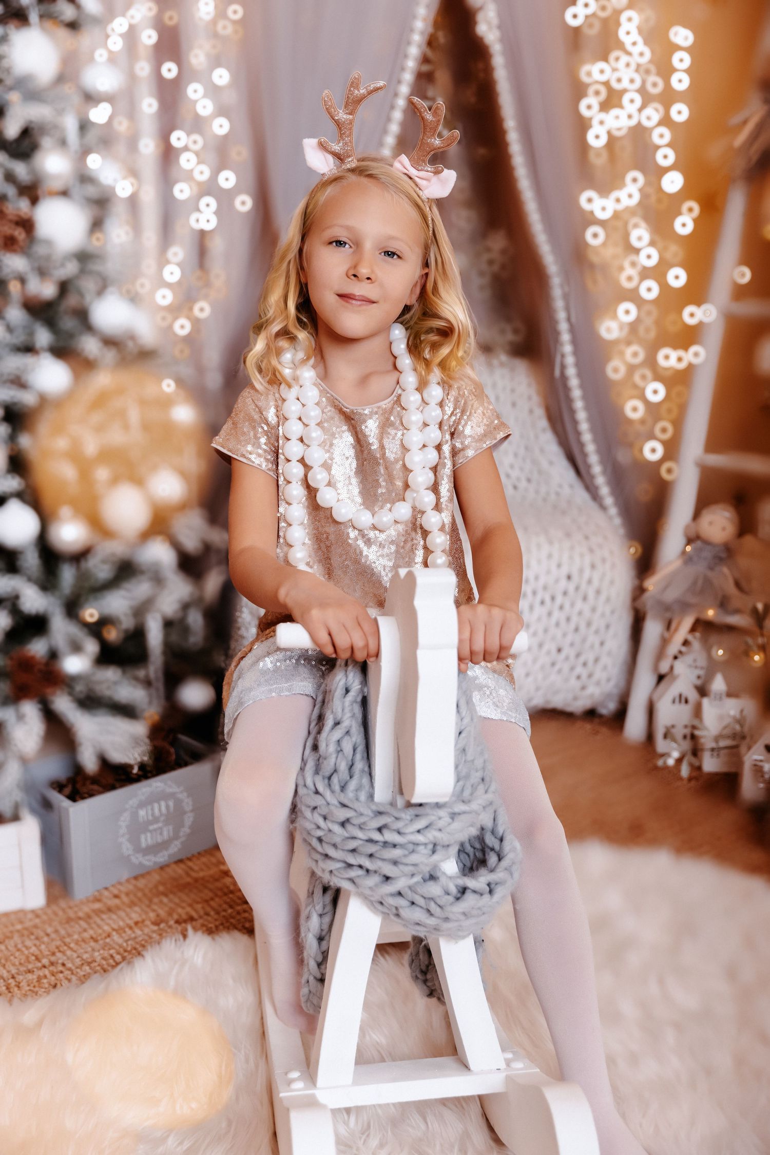 A young blonde girl in a sequin dress with reindeer antlers sits on a white rocking horse in a Christmas setting.