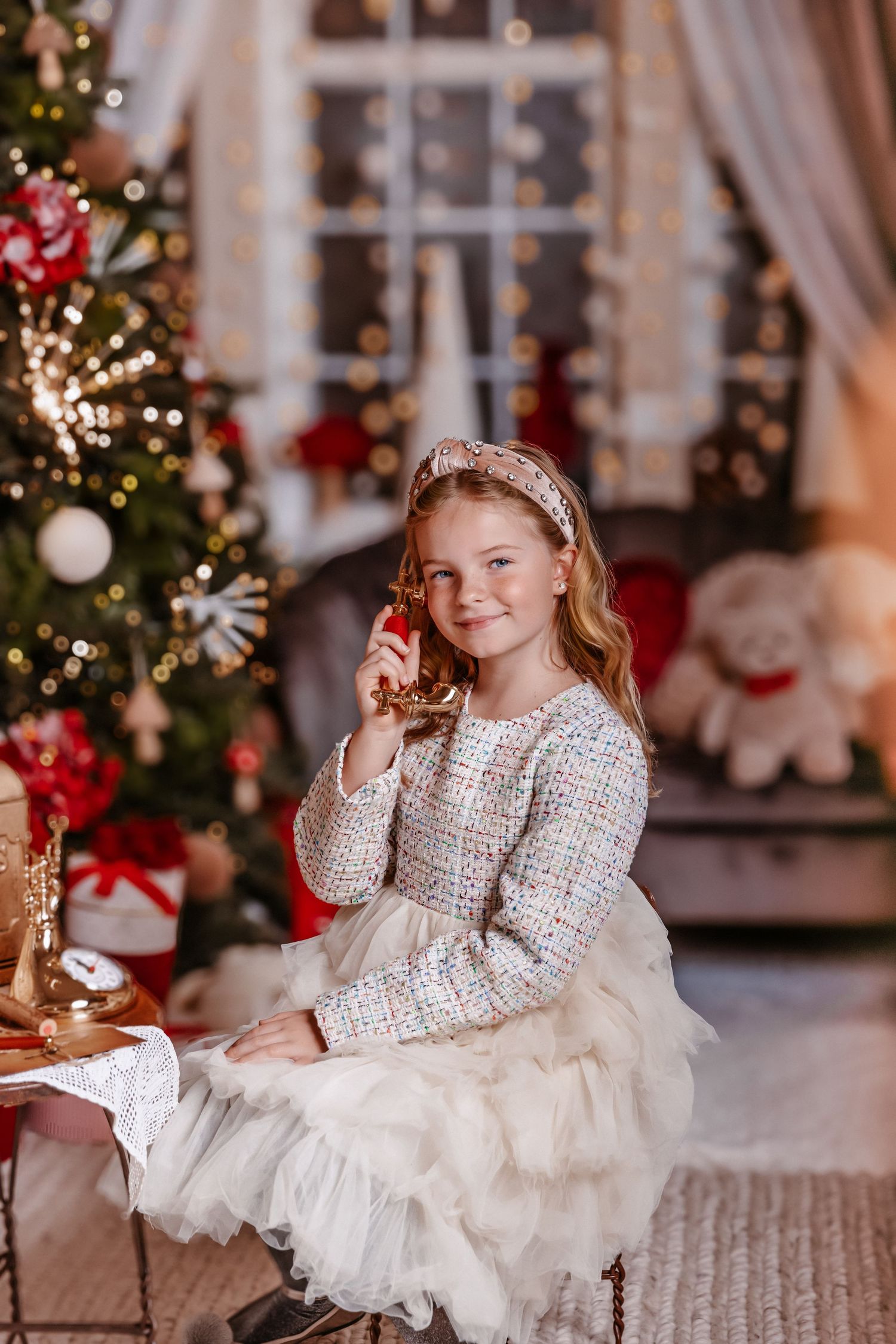 Smiling girl in tweed outfit sits by Christmas tree holding ornament in warmly lit holiday setting.