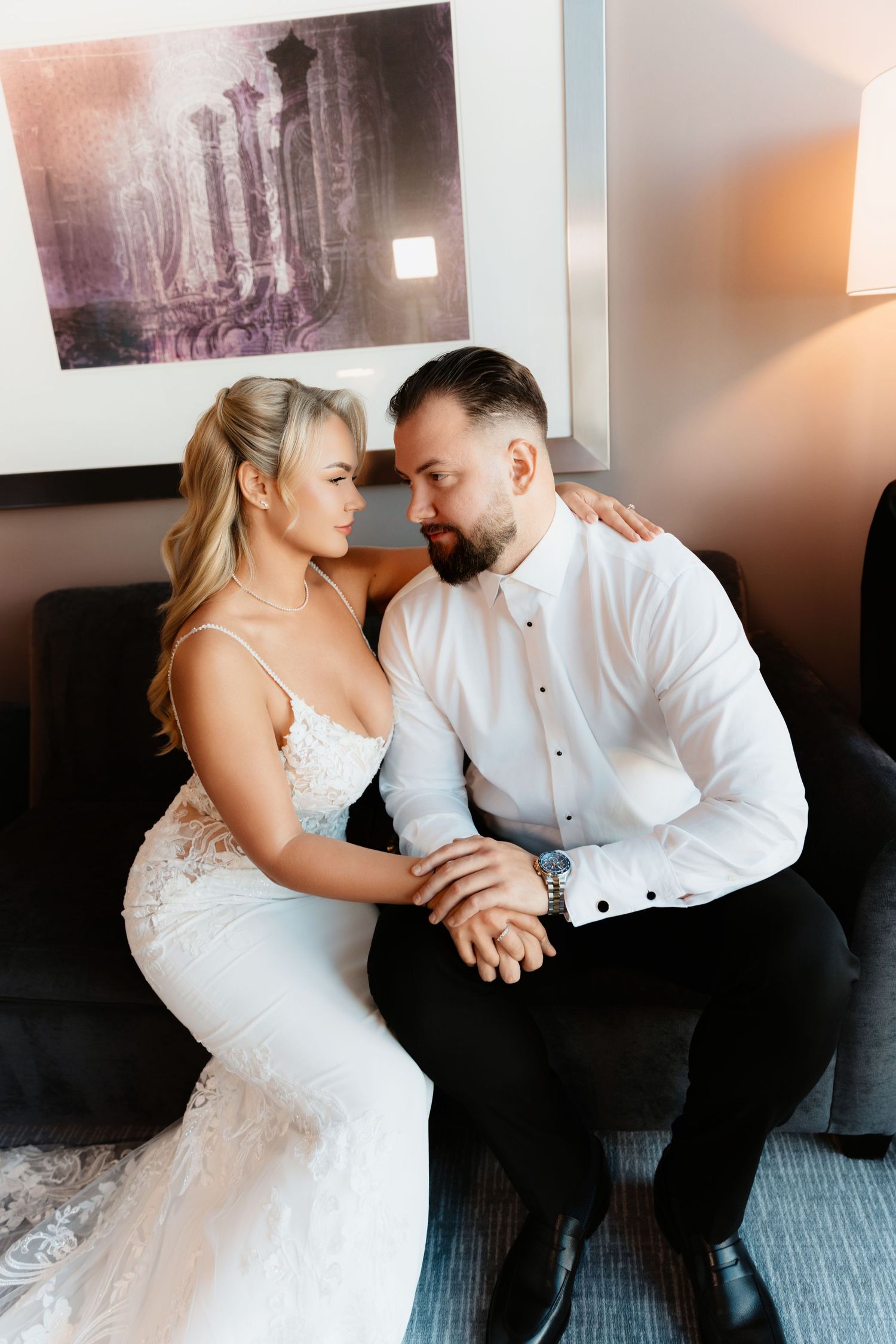 Bride in lace gown and bearded groom gaze at each other while seated on hotel sofa.