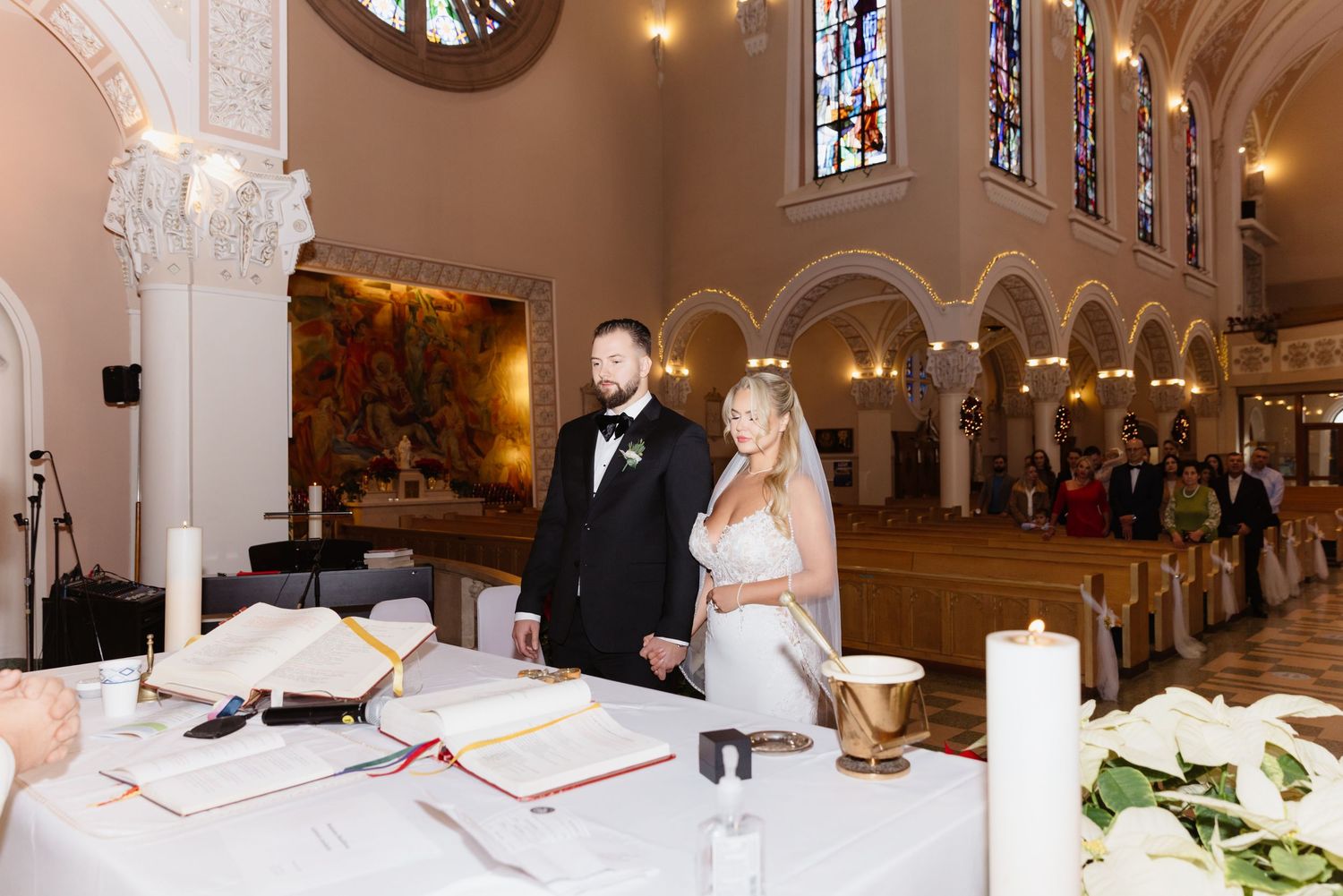 Bride and groom stand at signing table inside beautiful church with stained glass windows and wedding guests seated.