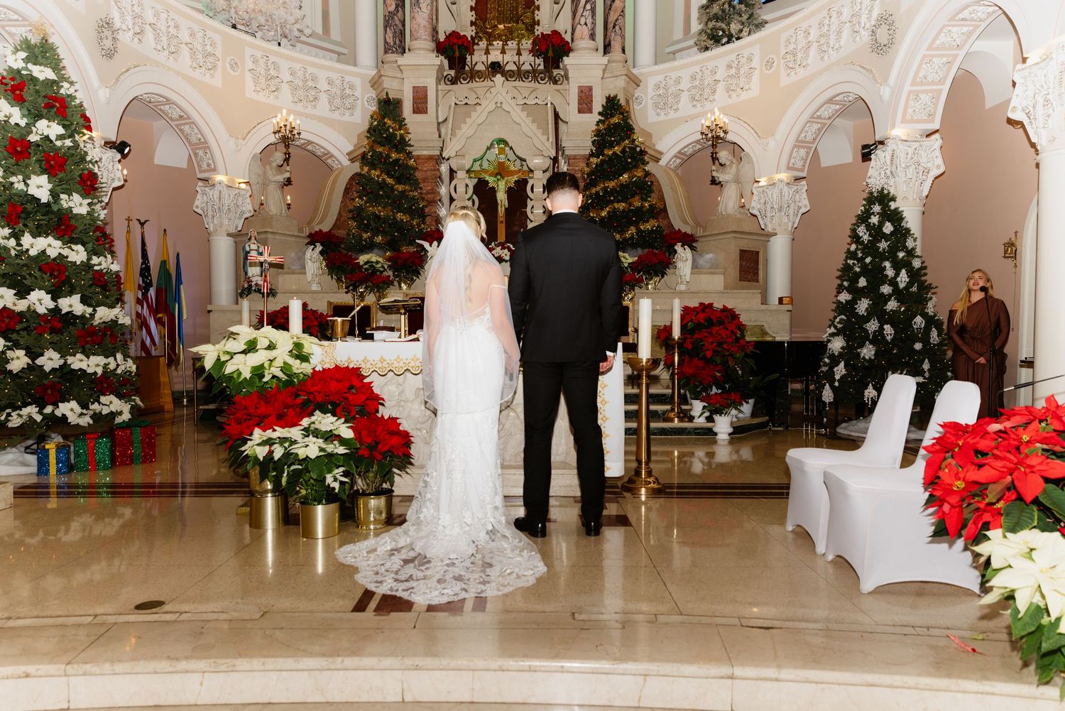 Bride and groom stand at altar surrounded by Christmas trees and poinsettias in decorated church.