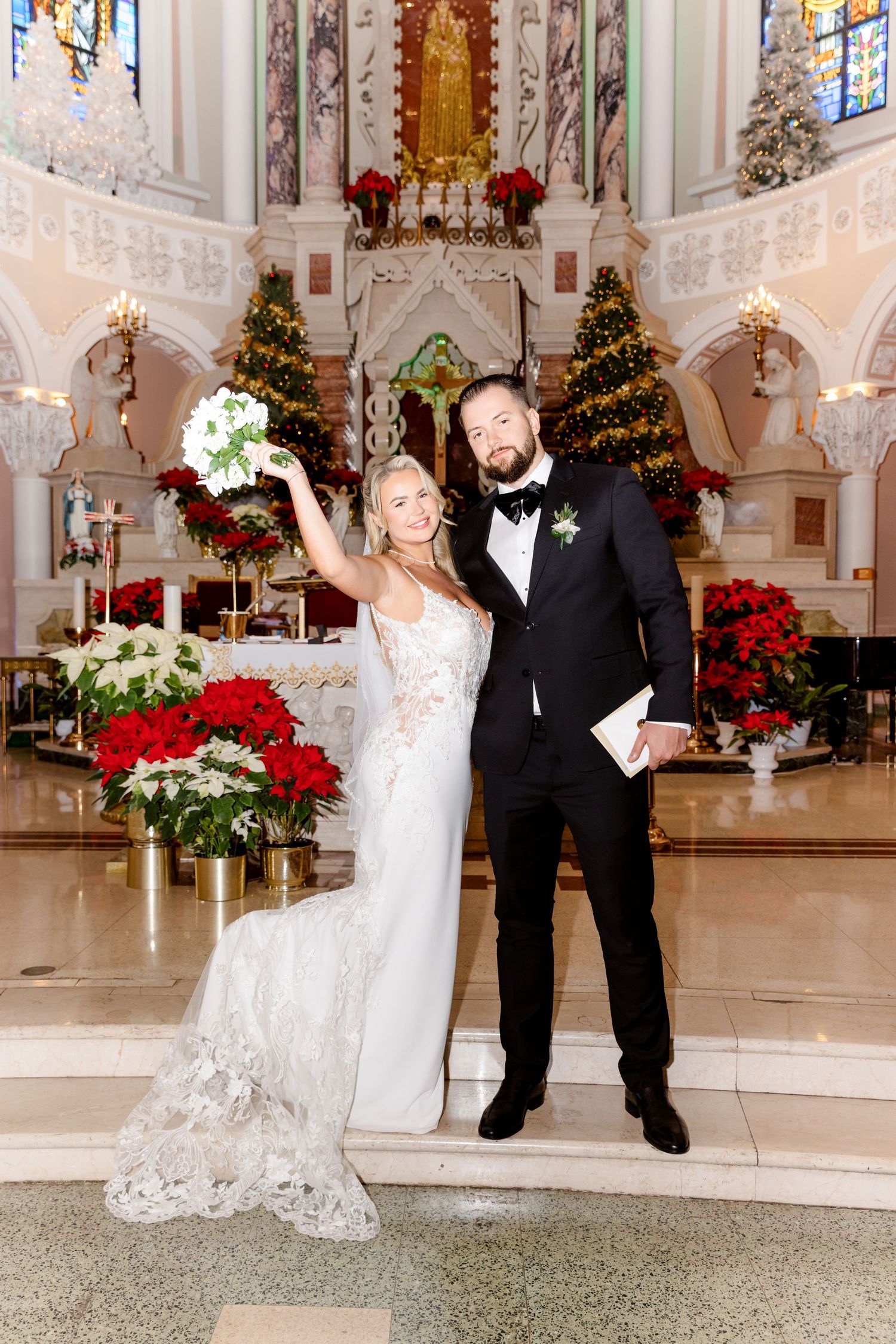 Bride and groom pose at church altar with Christmas decorations, poinsettias, and trees in background.