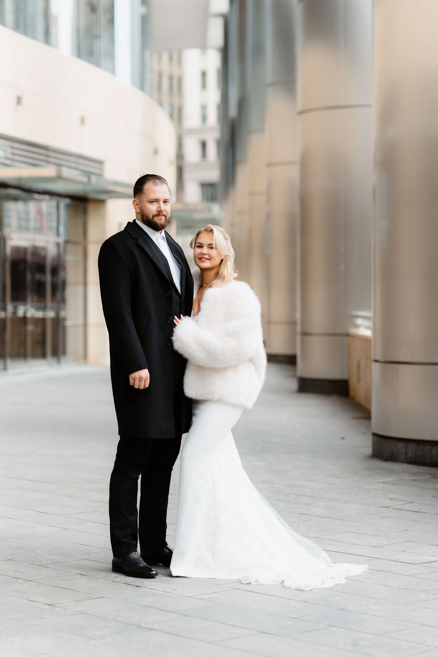 Bride wearing white fur stole and groom in black tuxedo pose together on city sidewalk.