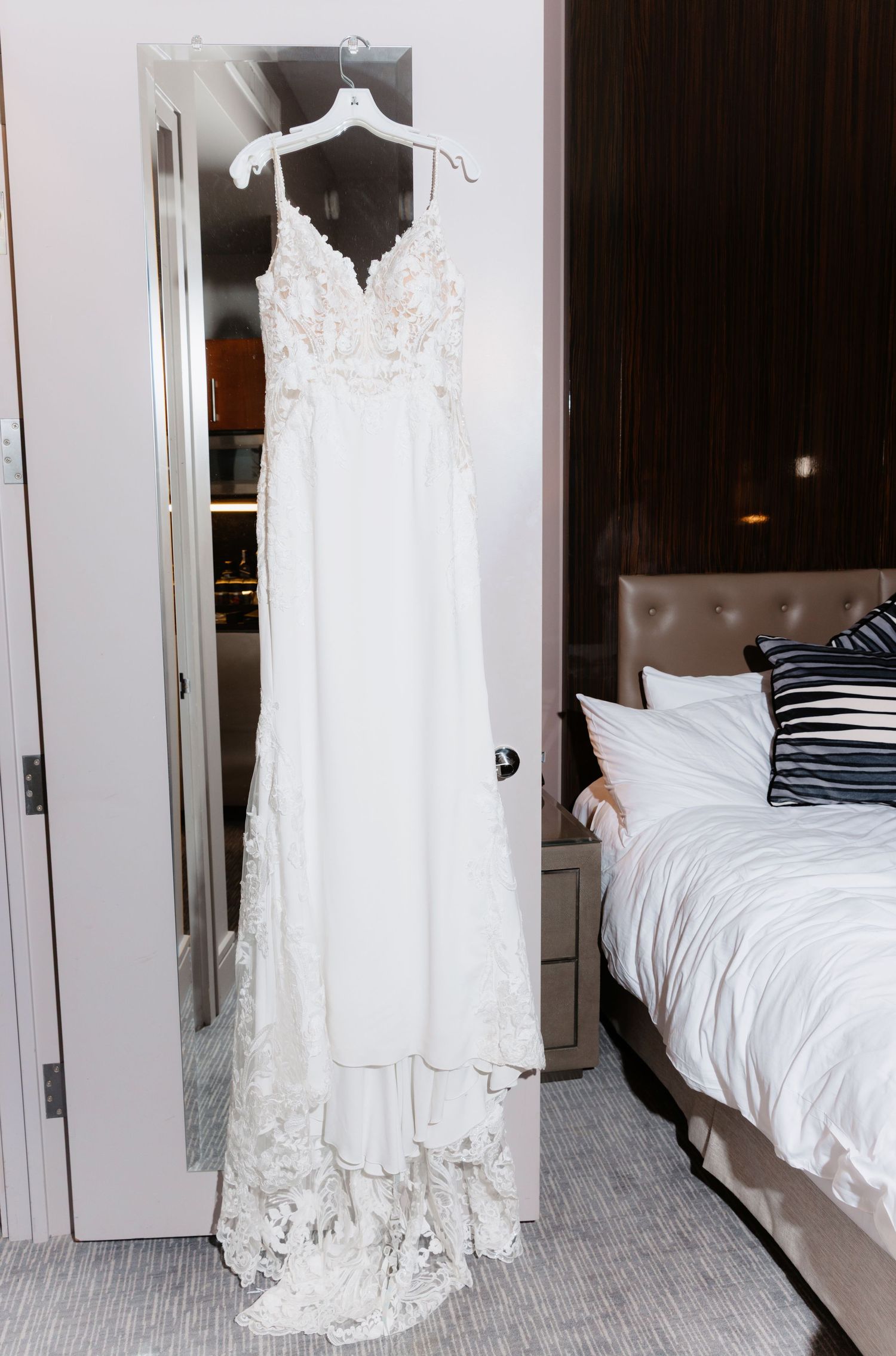 Elegant lace and chiffon wedding gown hanging on door in hotel room with city view window.
