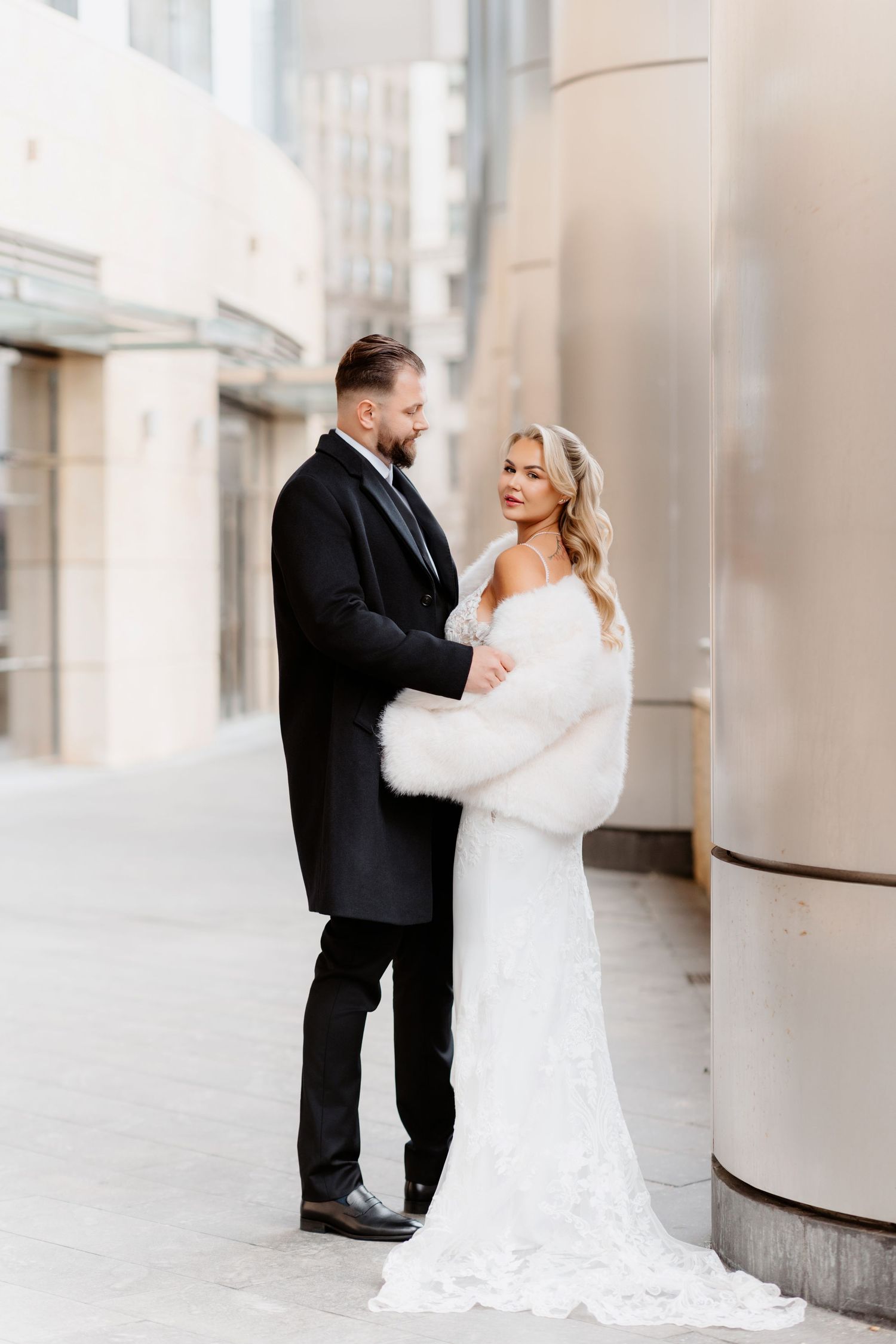 Elegant bride in lace gown and white fur wrap stands with bearded groom in black suit outdoors.