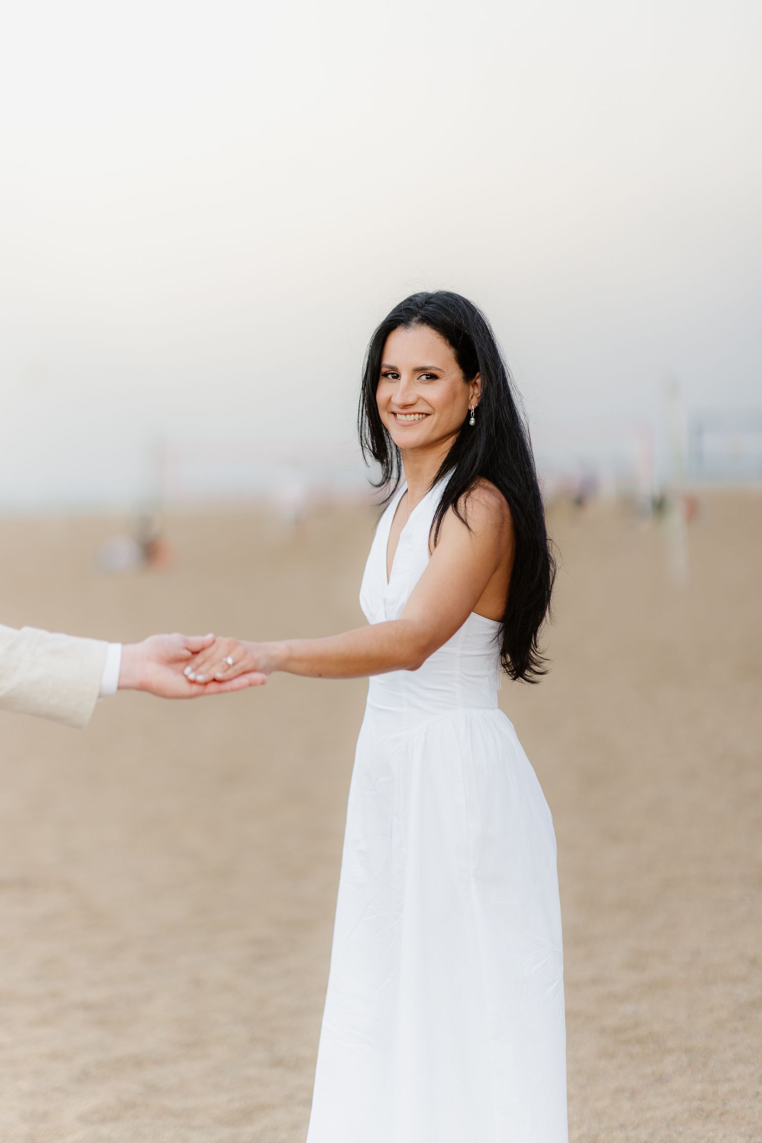 A smiling dark-haired woman in a white dress holds hands with her partner on a sandy beach.