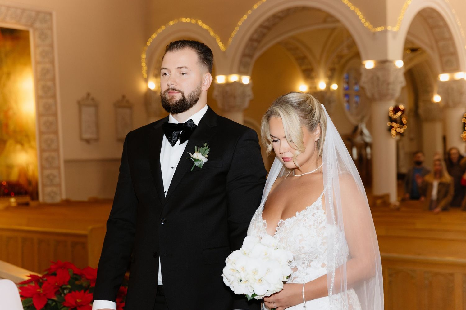 Bride in lace gown holding white bouquet stands beside groom in tuxedo during church wedding ceremony.