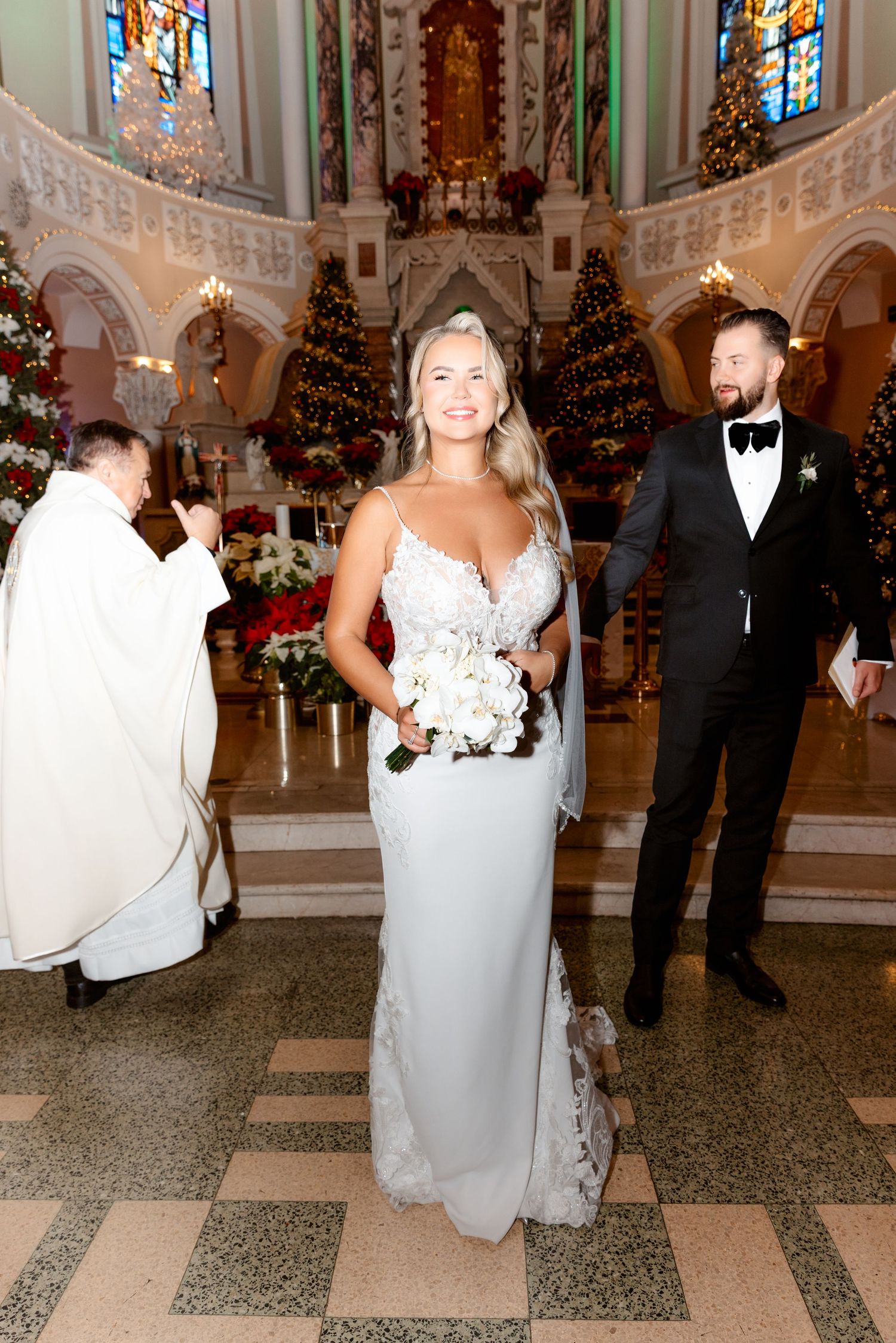 Bride smiling holding white bouquet at church ceremony with groom and priest visible in background.