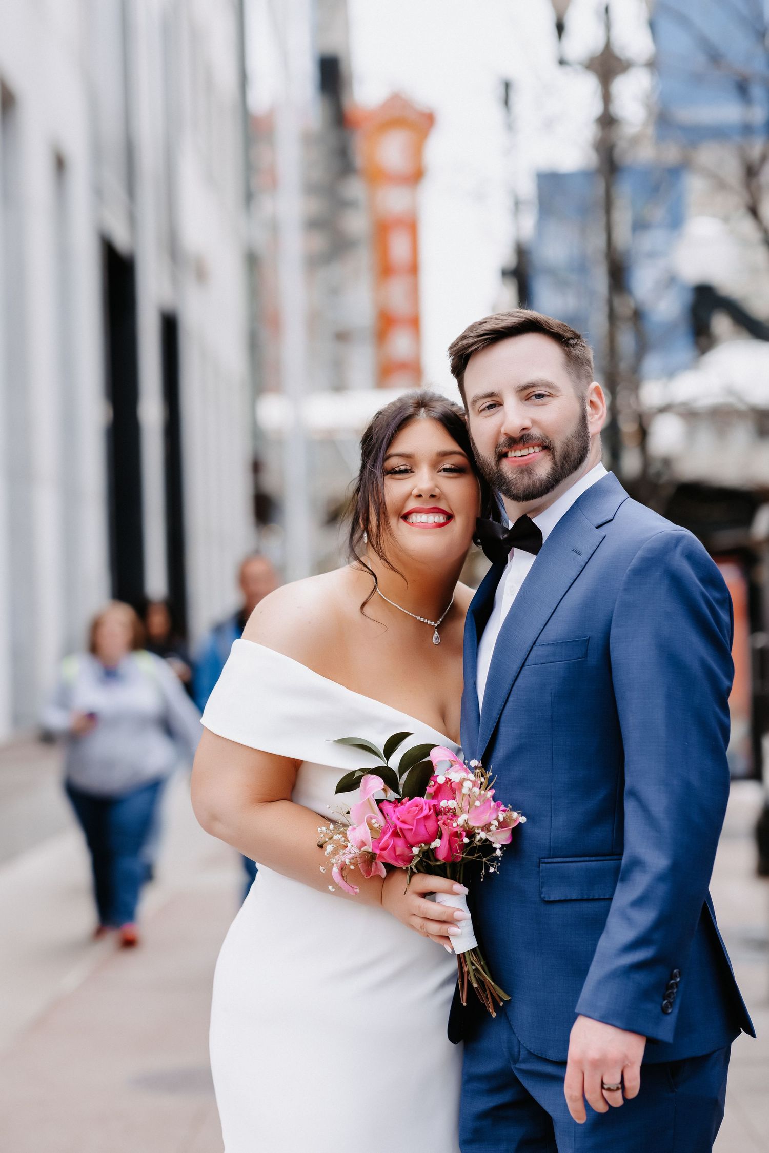 Smiling wedding couple poses on Chicago street, bride in off-shoulder gown holding pink bouquet, groom in navy suit.