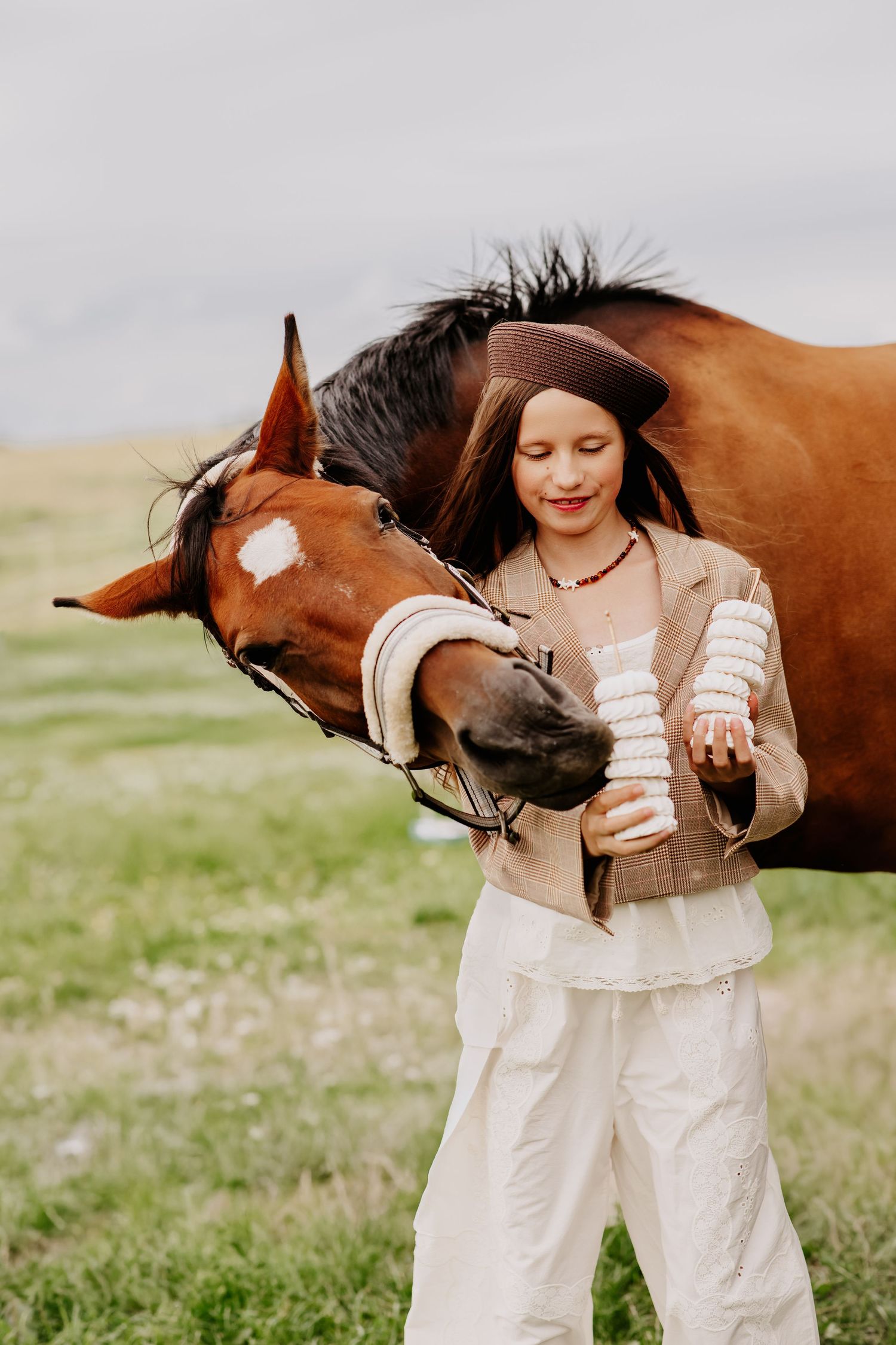 Girl in brown beret and beige outfit feeding a chestnut horse white stacked treats in an open green field.