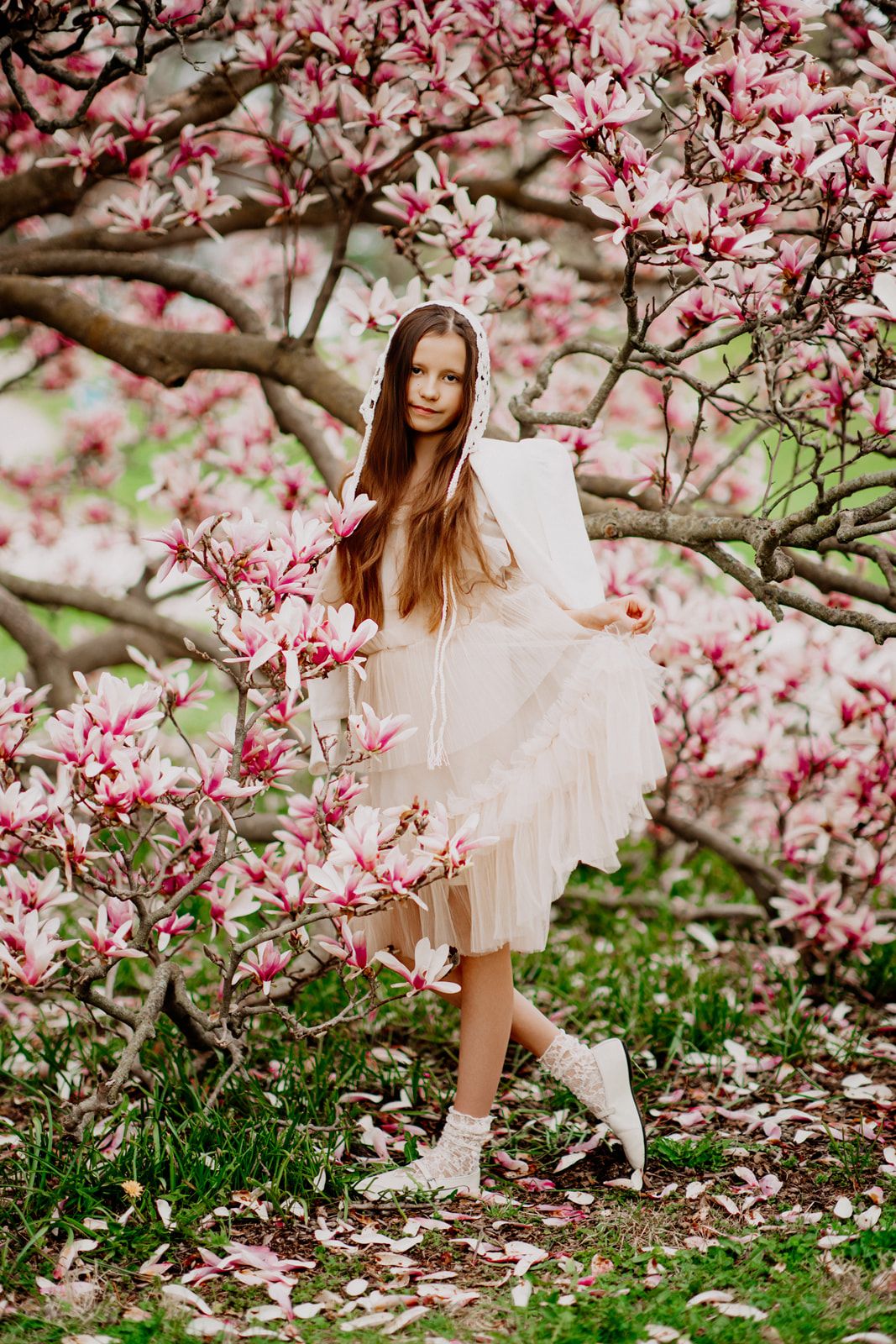 Young girl in white dress standing among blooming pink magnolia trees in spring garden.
