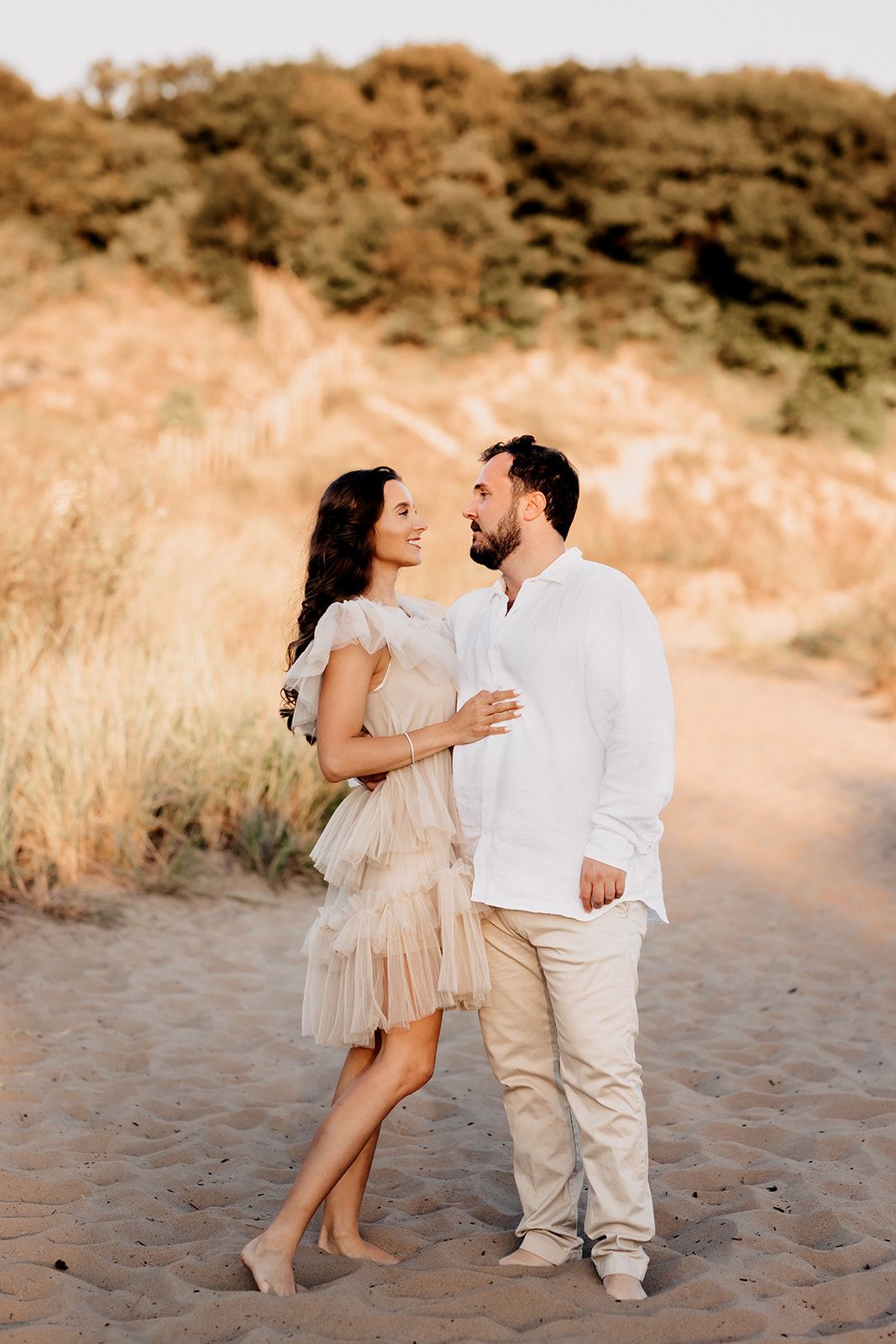 Romantic couple in beige and white outfits gazing at each other on a sandy beach at sunset.