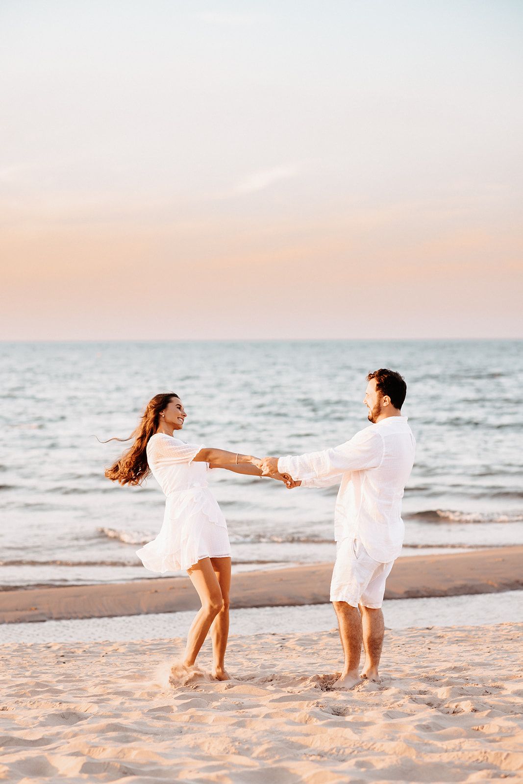 Happy couple in white outfits dancing on a sandy beach at sunset with ocean waves in the background.