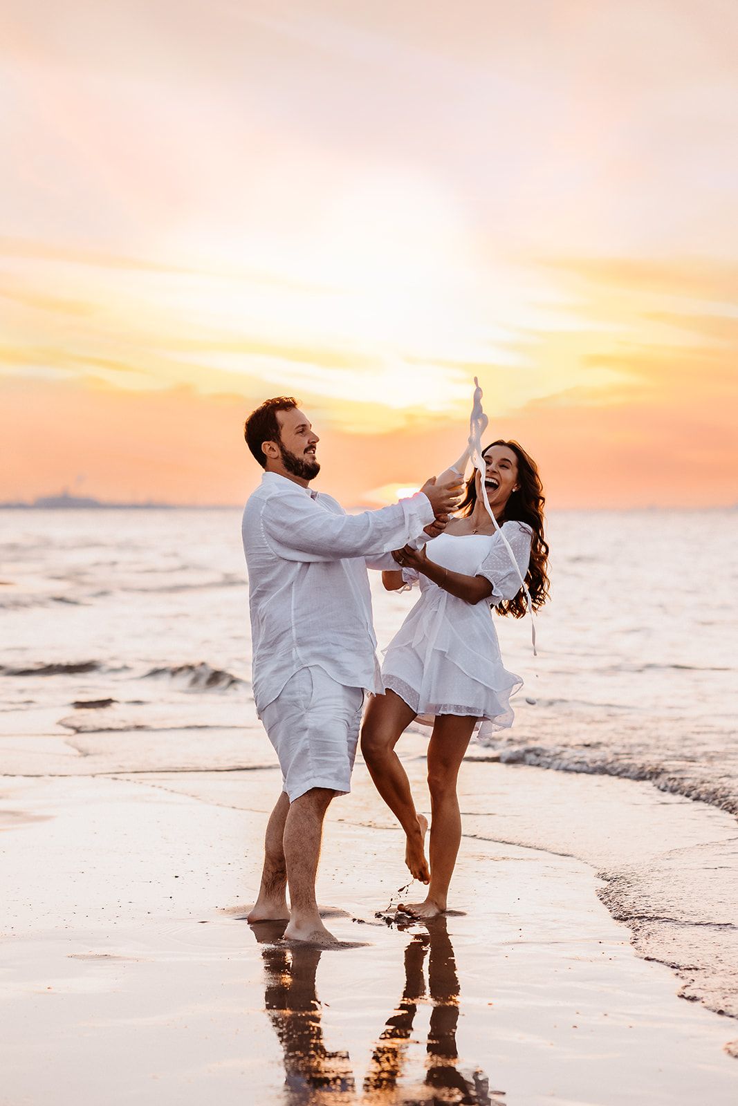 Happy couple in white outfits celebrating with champagne on a beach at golden sunset.