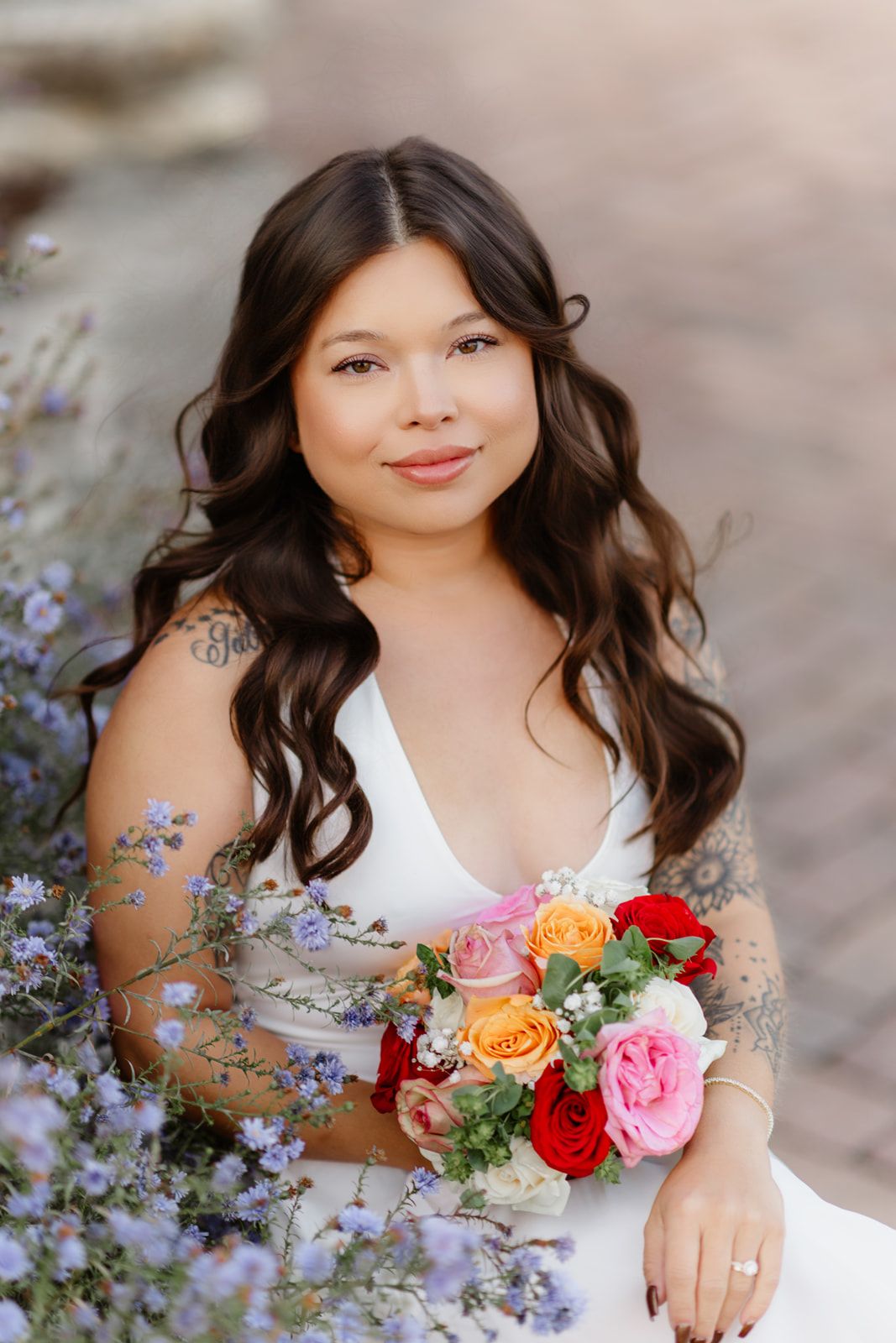 Bride in white dress holding colorful rose bouquet, sitting among purple wildflowers with tattoos visible on her arm.