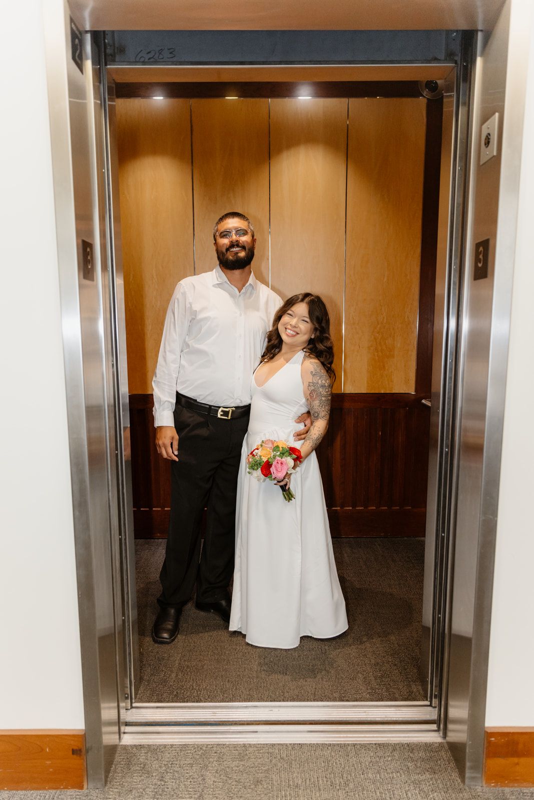 Newlywed couple in wedding attire smiling together inside an elevator, bride holding colorful bouquet.