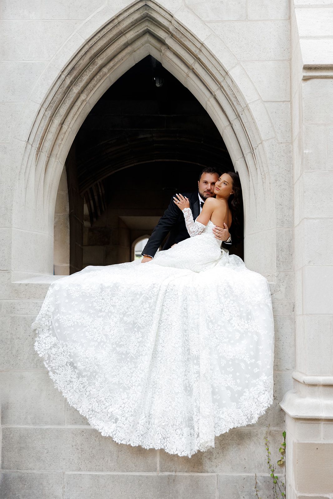 Groom holds bride in a stunning lace wedding dress while sitting in a Gothic stone archway entrance.