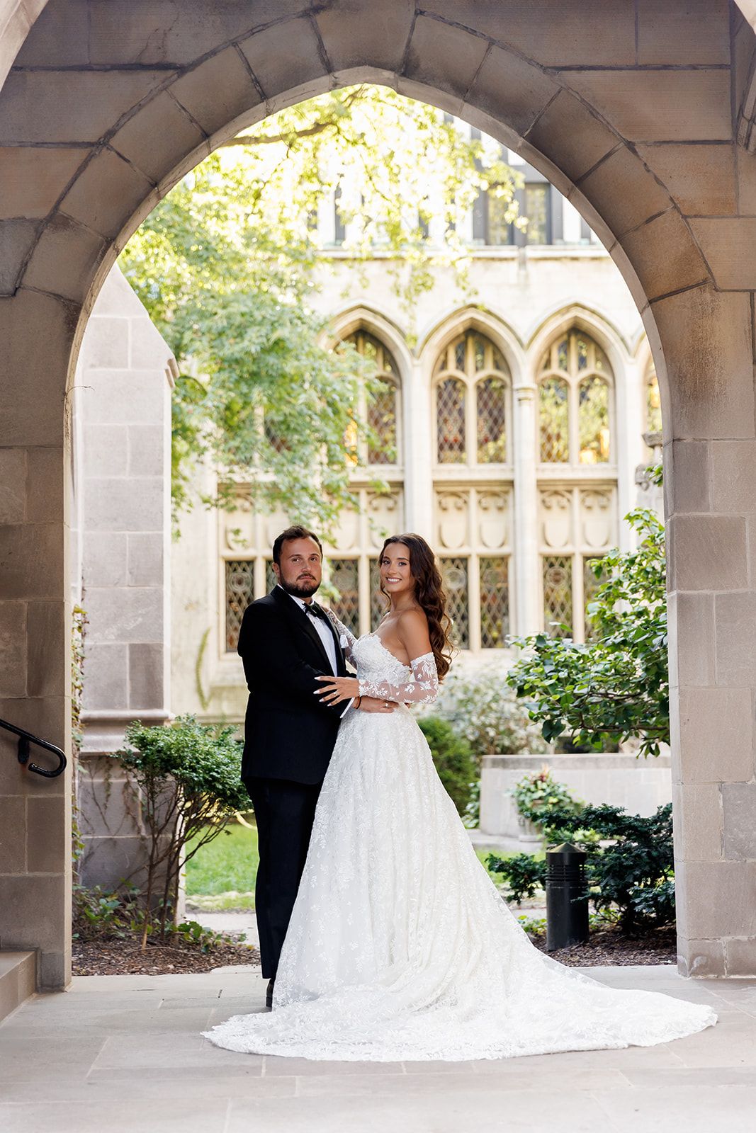 Elegant couple in wedding attire standing under a stone archway with Gothic windows and lush greenery backdrop.