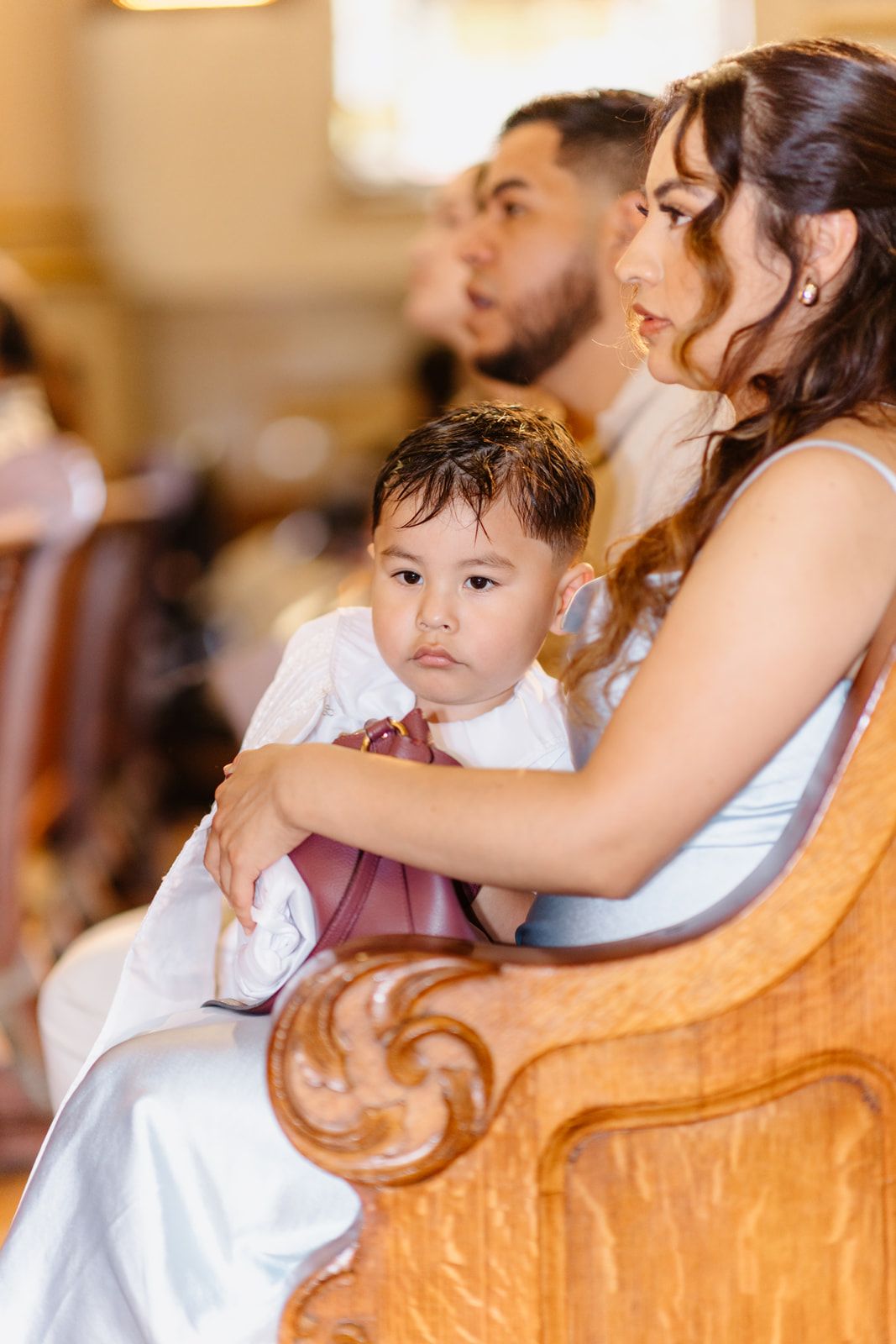 Young boy in white sits on woman's lap in church pew during a religious baptism ceremony.