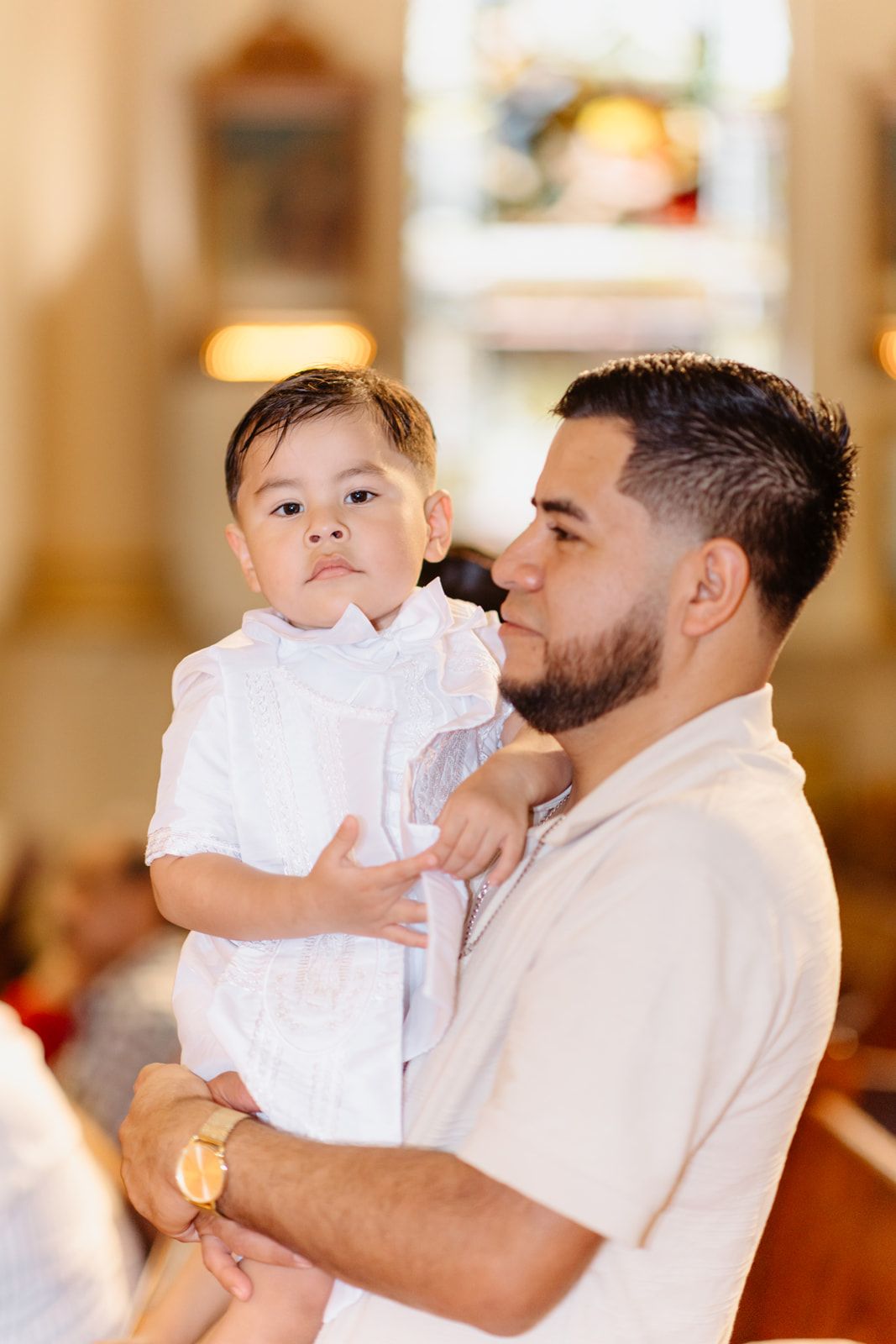 A bearded father lovingly holds his infant son dressed in white christening attire inside a church.
