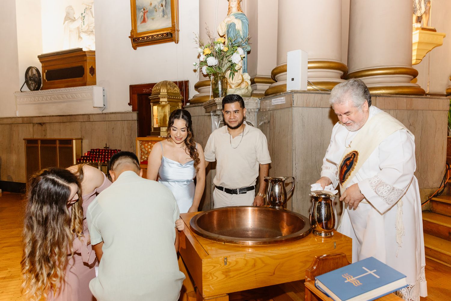 Priest performs baptism ceremony at copper font with family gathered around in a beautiful church.