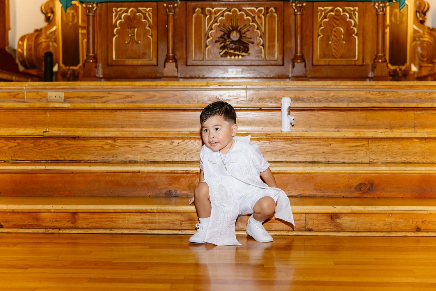 Adorable baby in white baptism outfit sitting on wooden church steps near ornate golden altar.